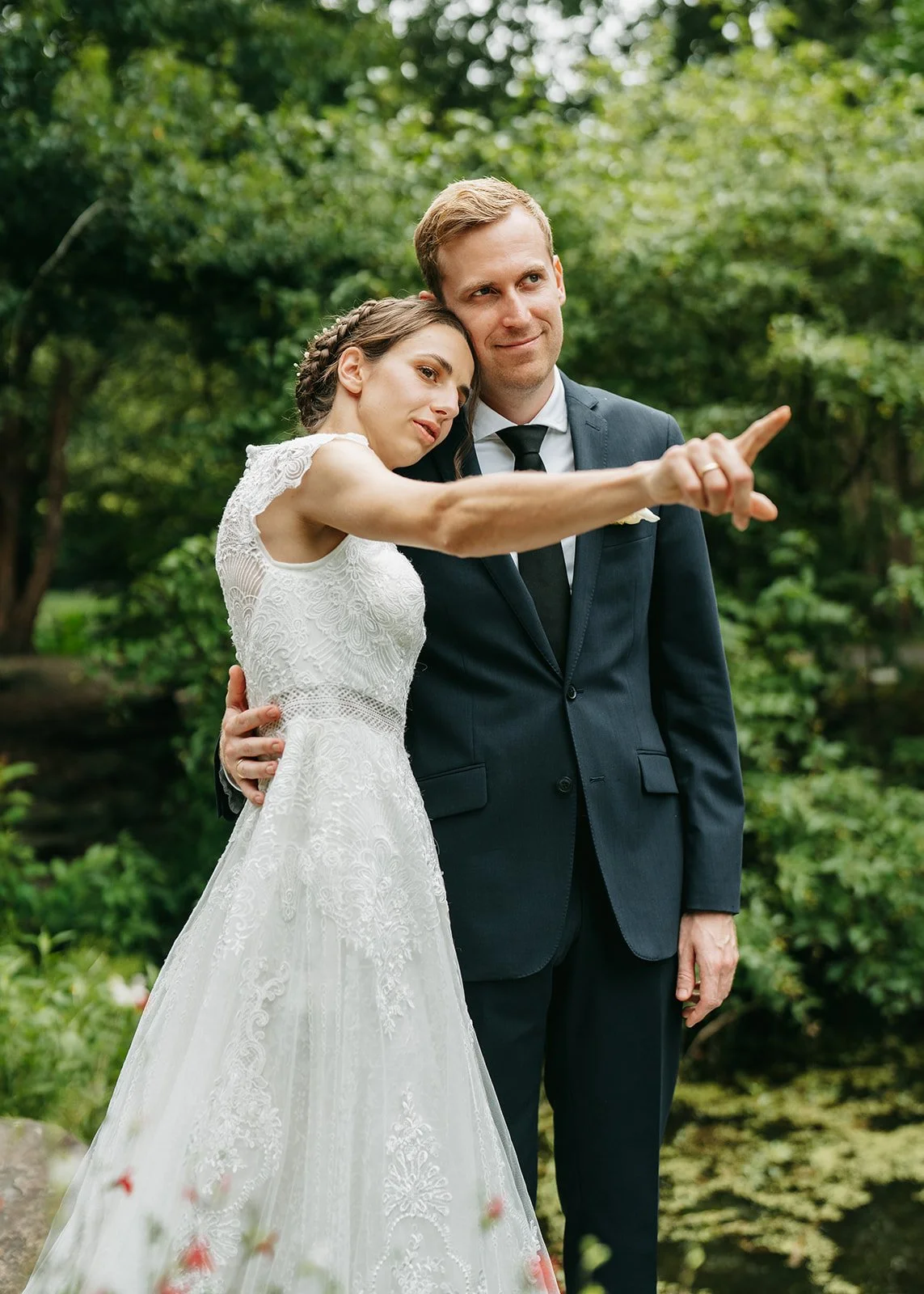 A bride and groom stand outdoors in a green setting. The bride, wearing a white lace wedding dress, leans her head on the groom's shoulder and points to something in the distance. The groom, dressed in a dark suit and tie, gently holds the bride arou