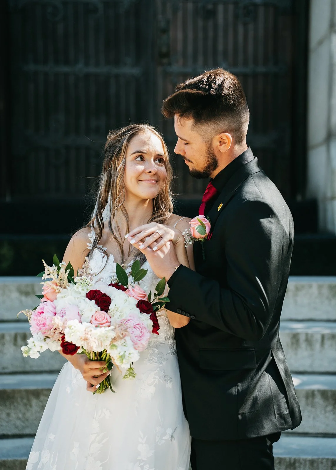 A bride and groom exchanging wedding vows outdoors. The bride is holding a bouquet of pink, white, and red flowers, and looking affectionately at the groom, who is holding her hands and wearing a black suit with a red tie and boutonniere.