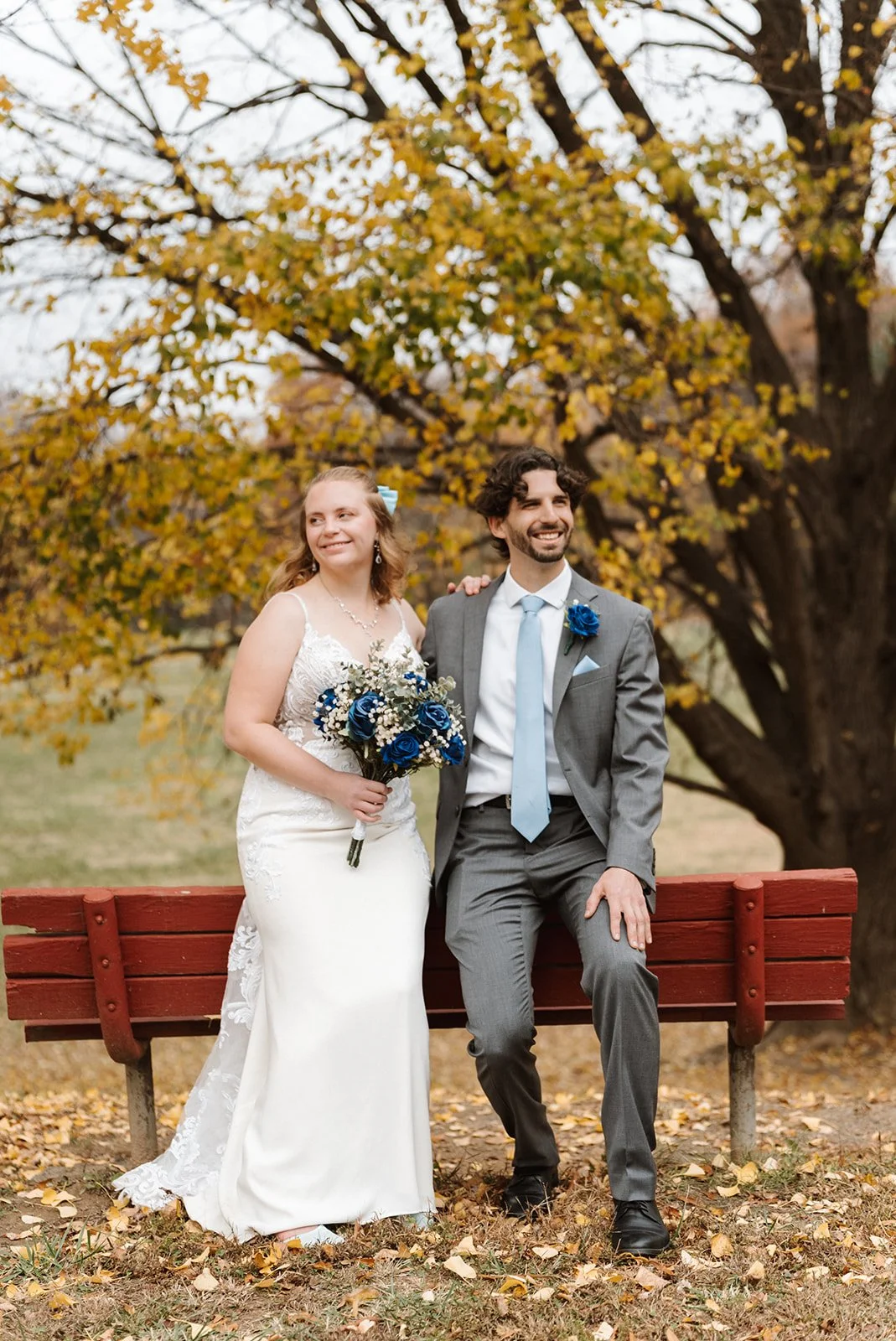 A bride and groom sit on a red park bench outdoors during autumn with yellow leaves in the background. The bride wears a white lace wedding dress and holds a bouquet of blue and white flowers. The groom wears a gray suit with a light blue tie and bou