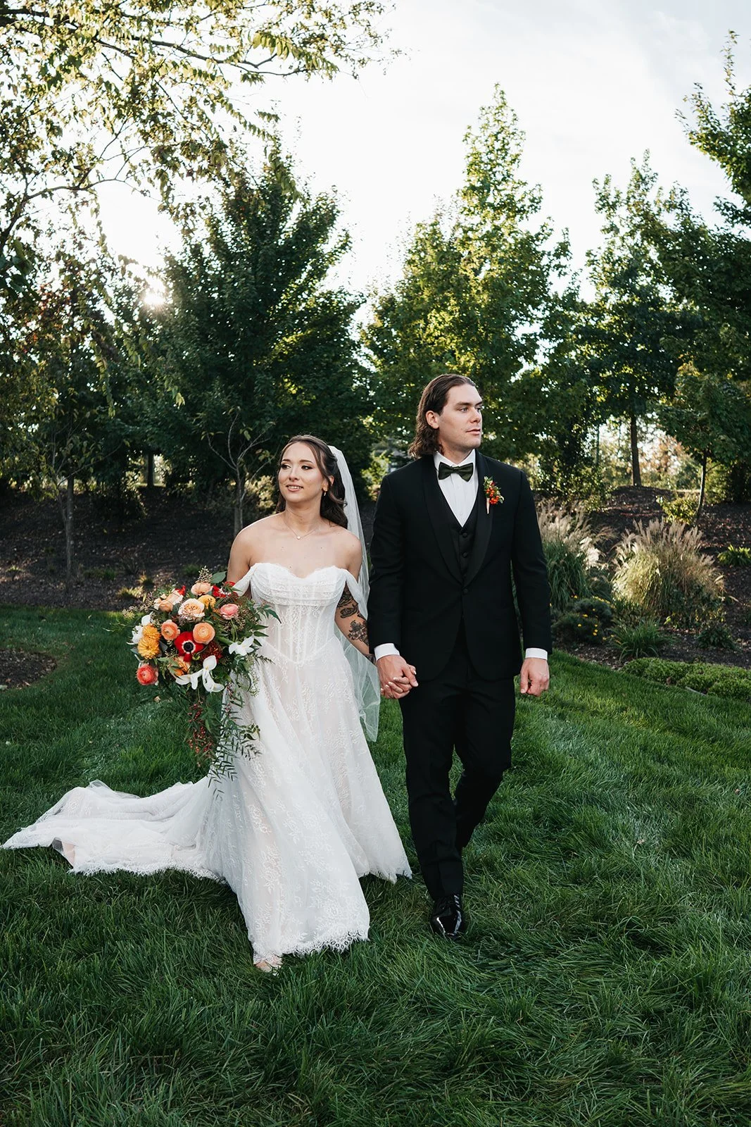 Bride and groom walking hand in hand outdoors during wedding, bride in white gown holding bouquet, groom in black tuxedo, lush green garden background.