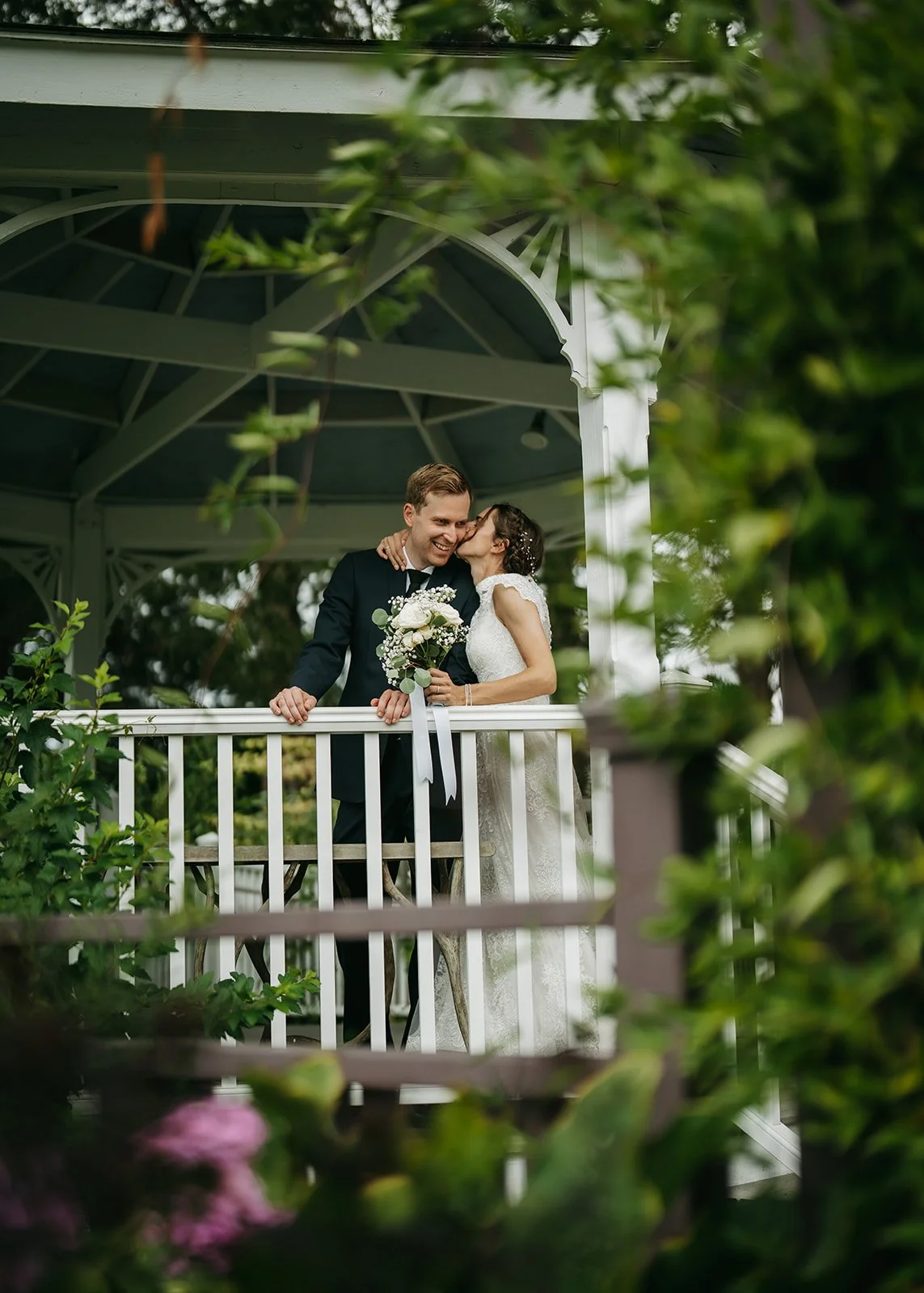 A bride and groom sharing a kiss on a gazebo, surrounded by greenery with pink flowers in the foreground.