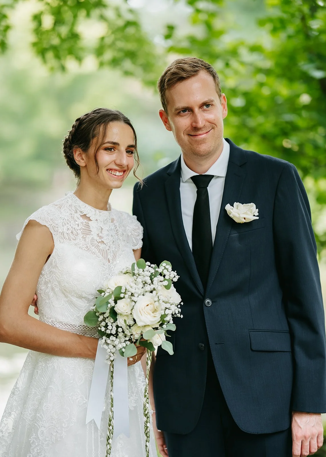 A bride in a white lace wedding dress holding a bouquet of white roses and greenery, standing next to a groom in a navy suit with a white boutonnière, outdoors with green trees in the background.