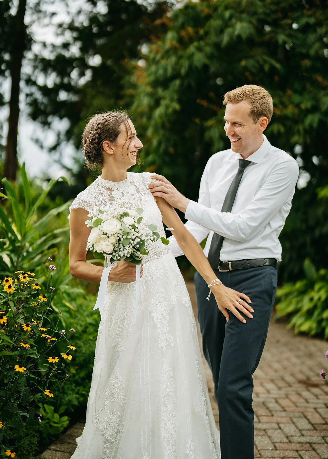 A bride in a white lace wedding dress holding a bouquet of white flowers standing outdoors, smiling at a man in a white shirt with a black tie, who is touching her shoulder, both surrounded by green foliage.