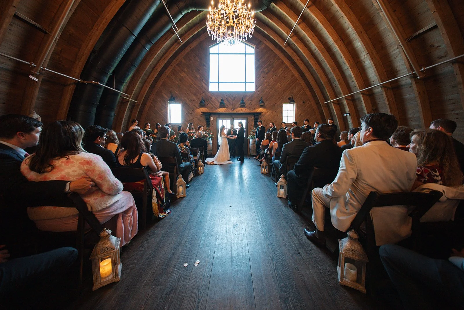 A wedding ceremony taking place inside a wooden arch-shaped venue with high curved ceilings and chandelier, where a couple is exchanging vows in front of an officiant, with guests seated on both sides.