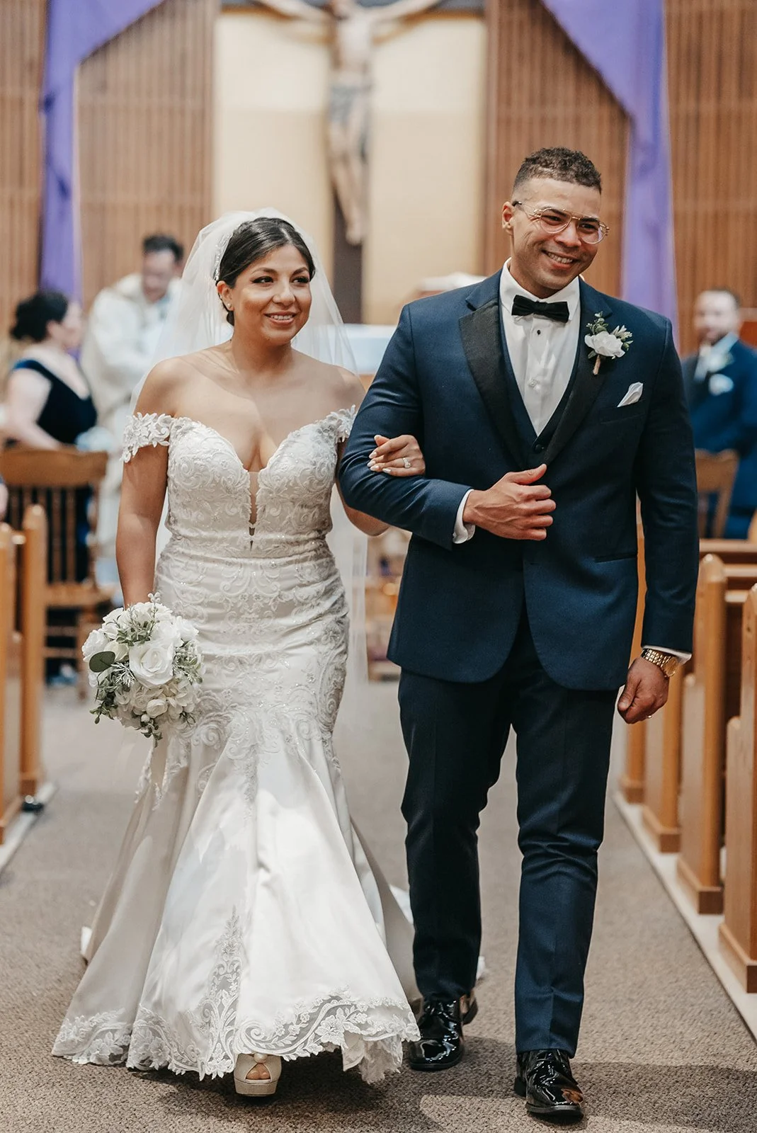 A bride and groom walking down the aisle after their wedding ceremony inside a church, with friends and family in the background.