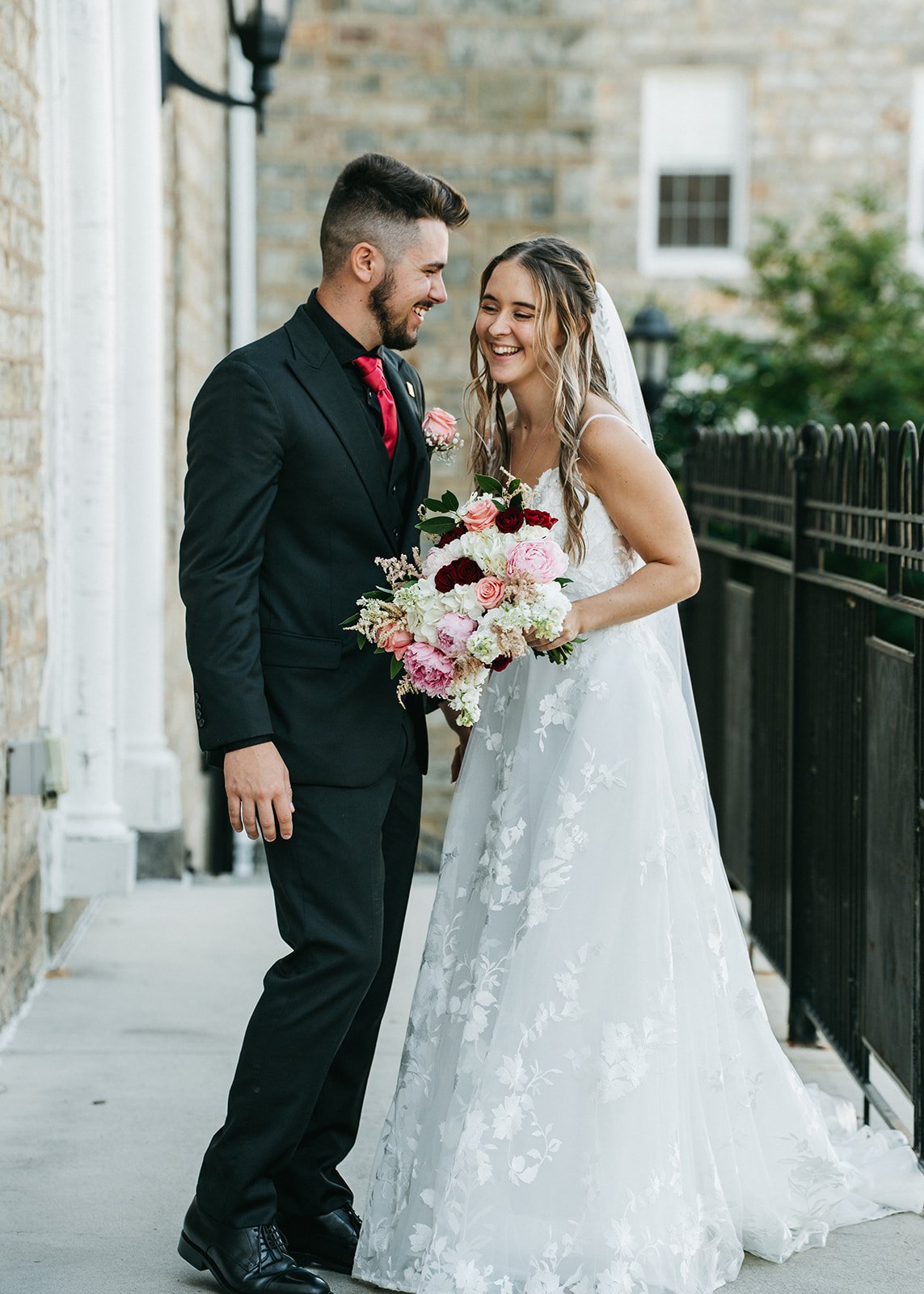 A newlywed couple stands together outdoors, smiling and looking at each other. The woman is wearing a white lace wedding gown and holding a bouquet of pink, white, and deep red flowers. The man is dressed in a black suit with a red tie and boutonnier