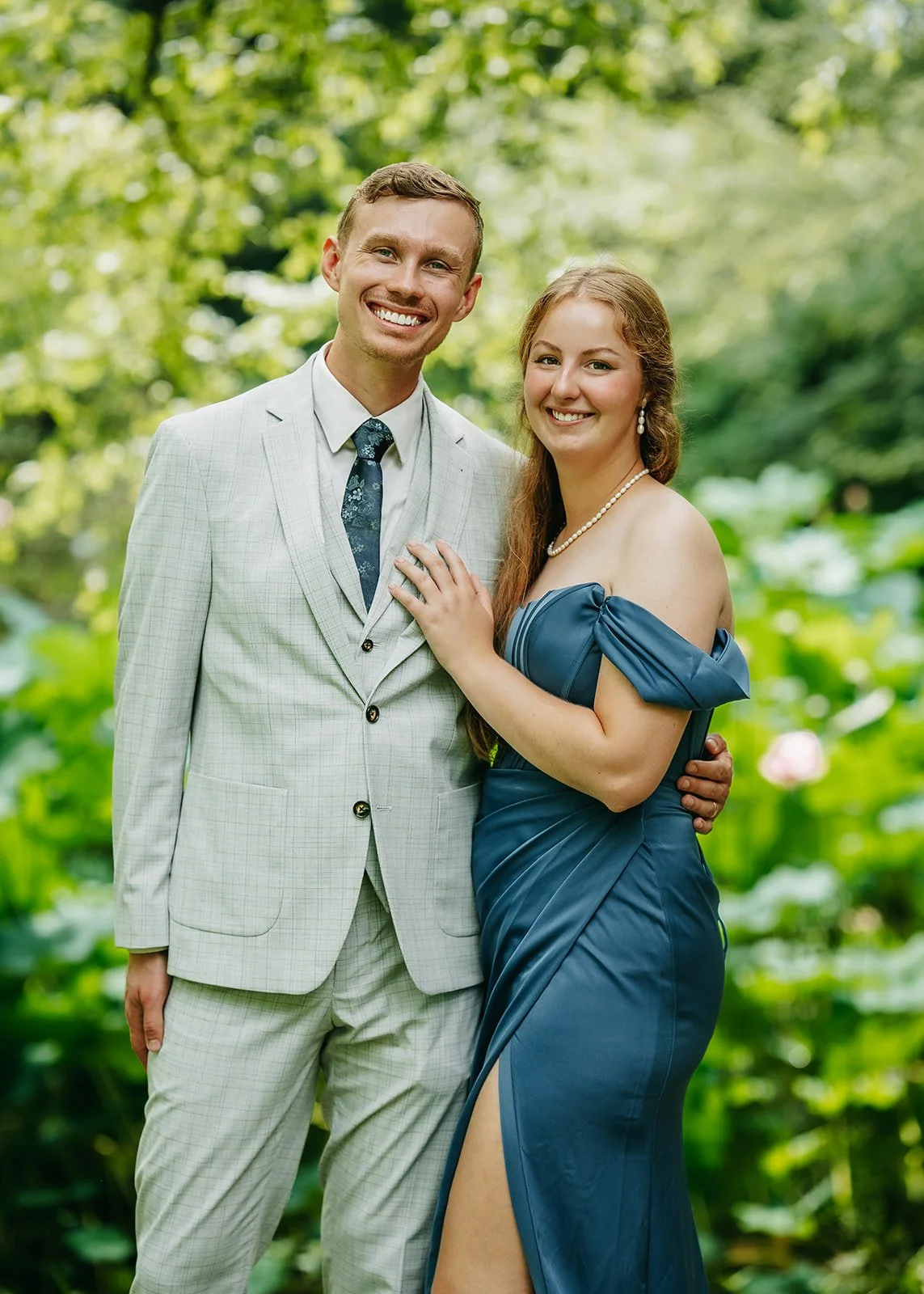 A young man and woman smiling outdoors in a lush green garden, dressed in formal attire. The man is wearing a light gray suit with a patterned tie, and the woman is in an off-the-shoulder blue gown with a pearl necklace. They are standing close toget