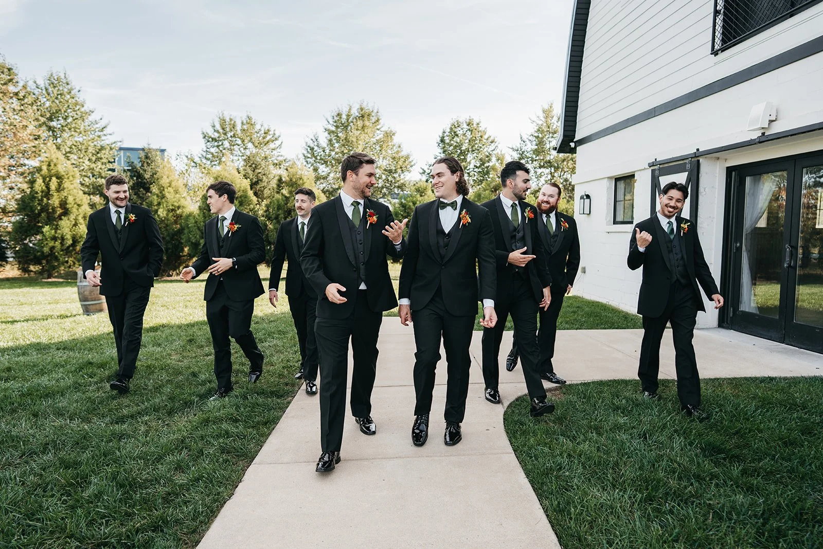 A group of eight men in black tuxedos and green ties walking outdoors on a sidewalk, smiling and conversing near a white building with greenery in the background.