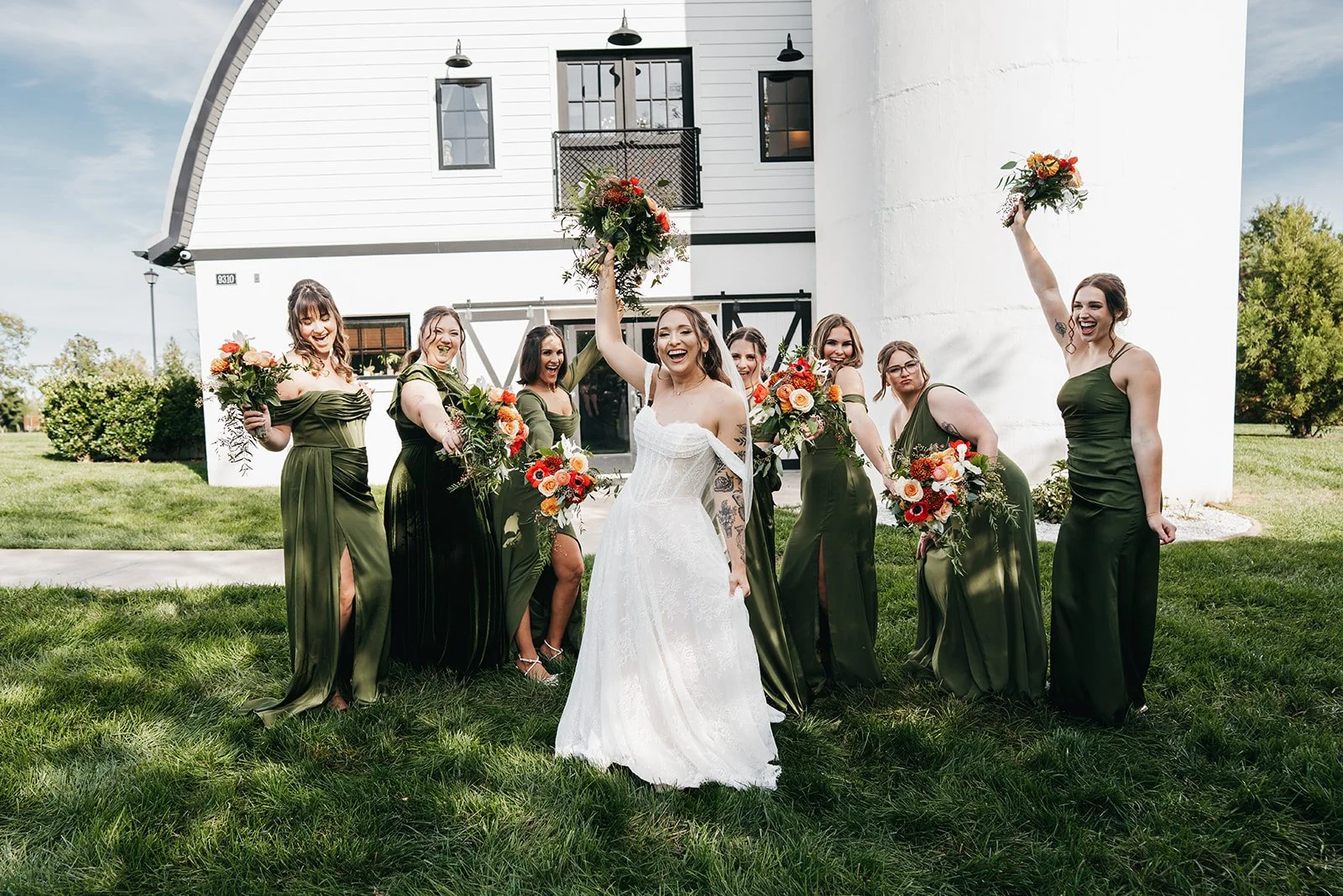 A bride in a white wedding dress surrounded by bridesmaids in matching green dresses, holding bouquets, posing outdoors on grass in front of a modern building with white exterior walls.