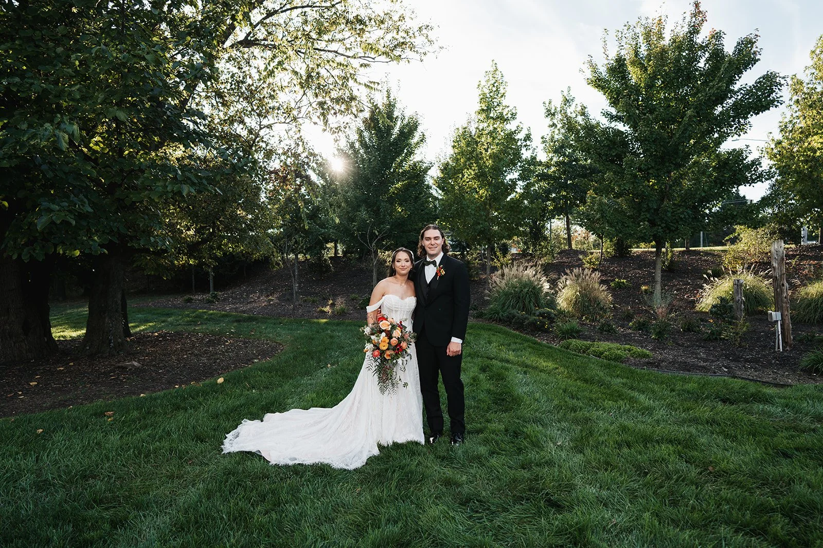 A bride and groom standing on grass in a park during their wedding. The bride is holding a bouquet and wearing a white wedding gown, while the groom is dressed in a black tuxedo with a bow tie. Trees and shrubbery surround them, and the sun is shinin