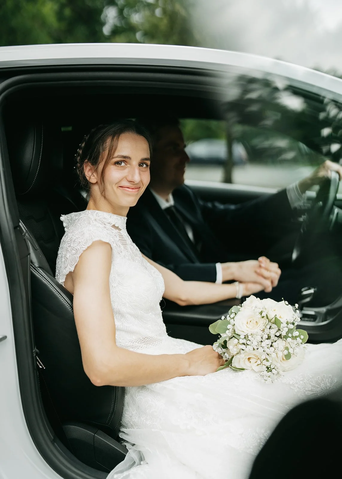 A bride sitting in the passenger seat of a car, holding a white floral bouquet, and smiling at the camera. The bride is wearing a lace wedding dress, and a man is driving the car, sitting in the driver's seat with his hands clasped. The background sh