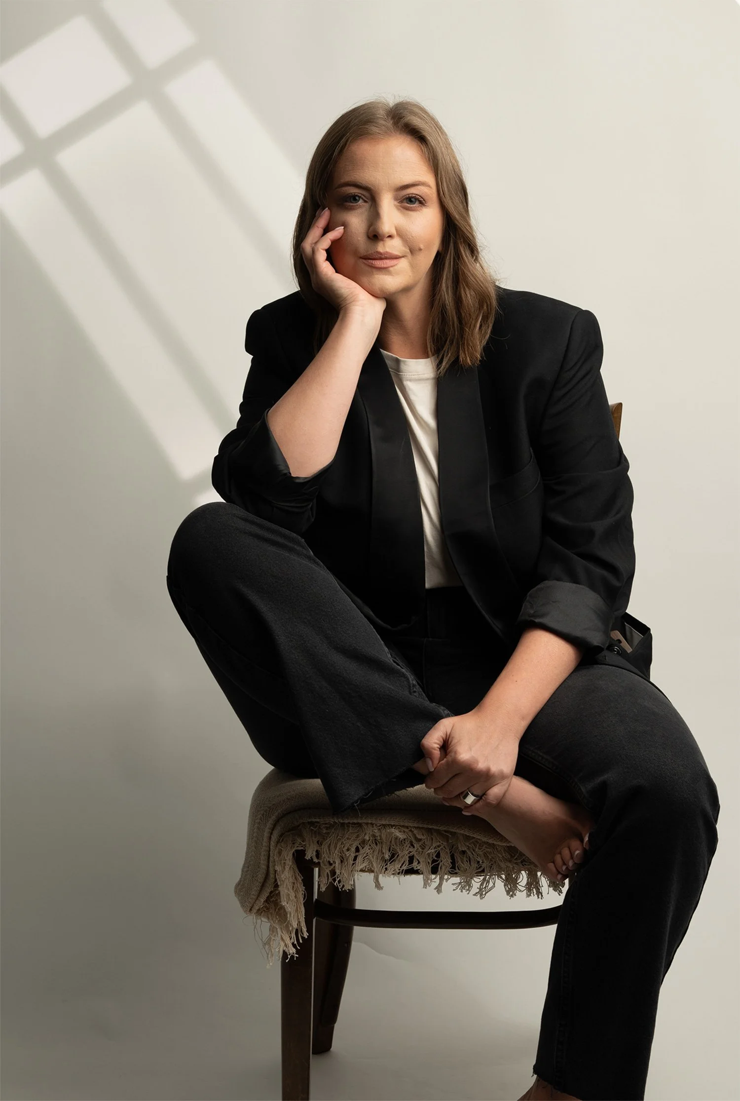 A woman with shoulder-length brown hair sitting on a wooden chair with a beige fringed cloth draped over it, resting her left hand on her cheek and her right ankle on her knee, against a plain light background with shadows cast on the wall.