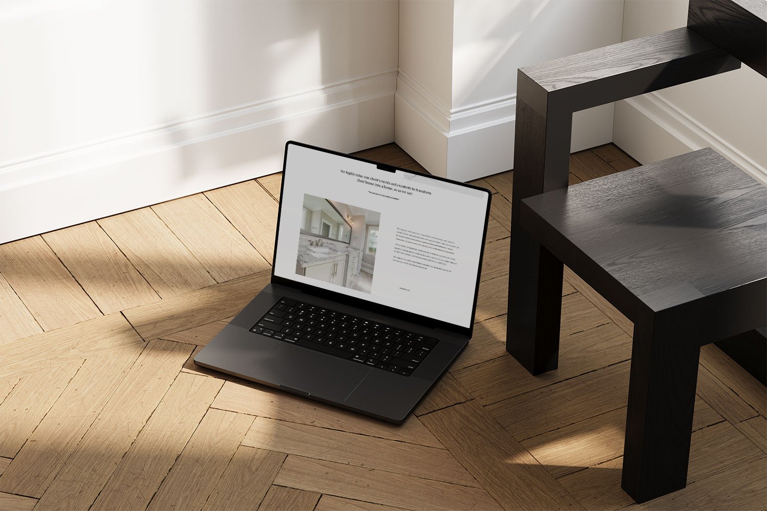 Open laptop on a wooden floor displaying a home renovation website, next to a black wooden table in a bright room with white walls and baseboards.
