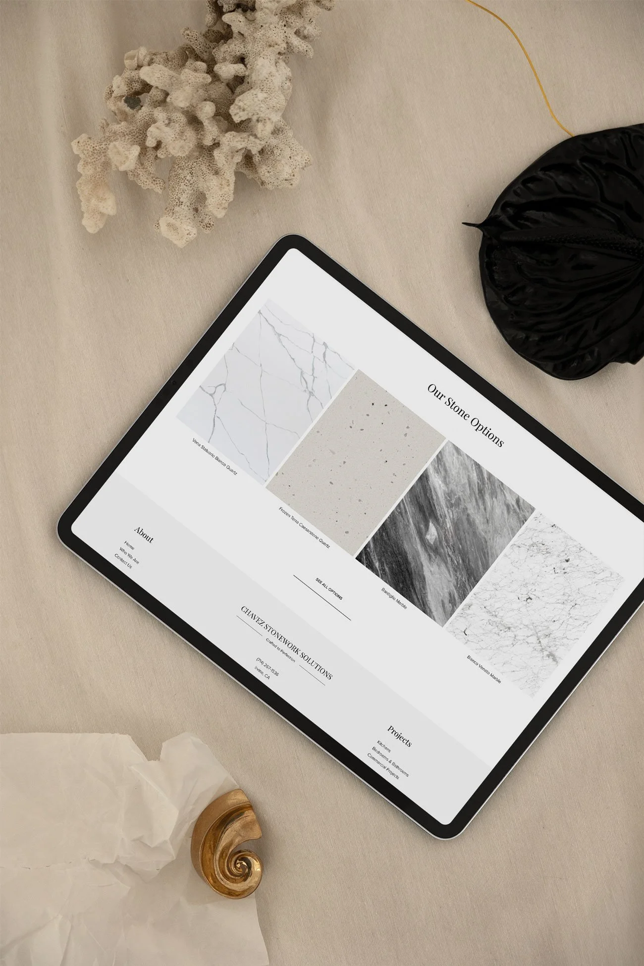 Tablet displaying stone options on a beige table, surrounded by coral, a black bag, and a seashell.