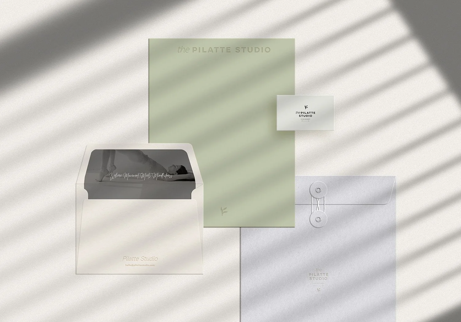Flat lay of Pilates Studio promotional materials, includes a white envelope, a flyer with a black and white photo, a green sheet of paper, a business card, and a white paper with a button clip, all casting soft shadows on a beige surface.
