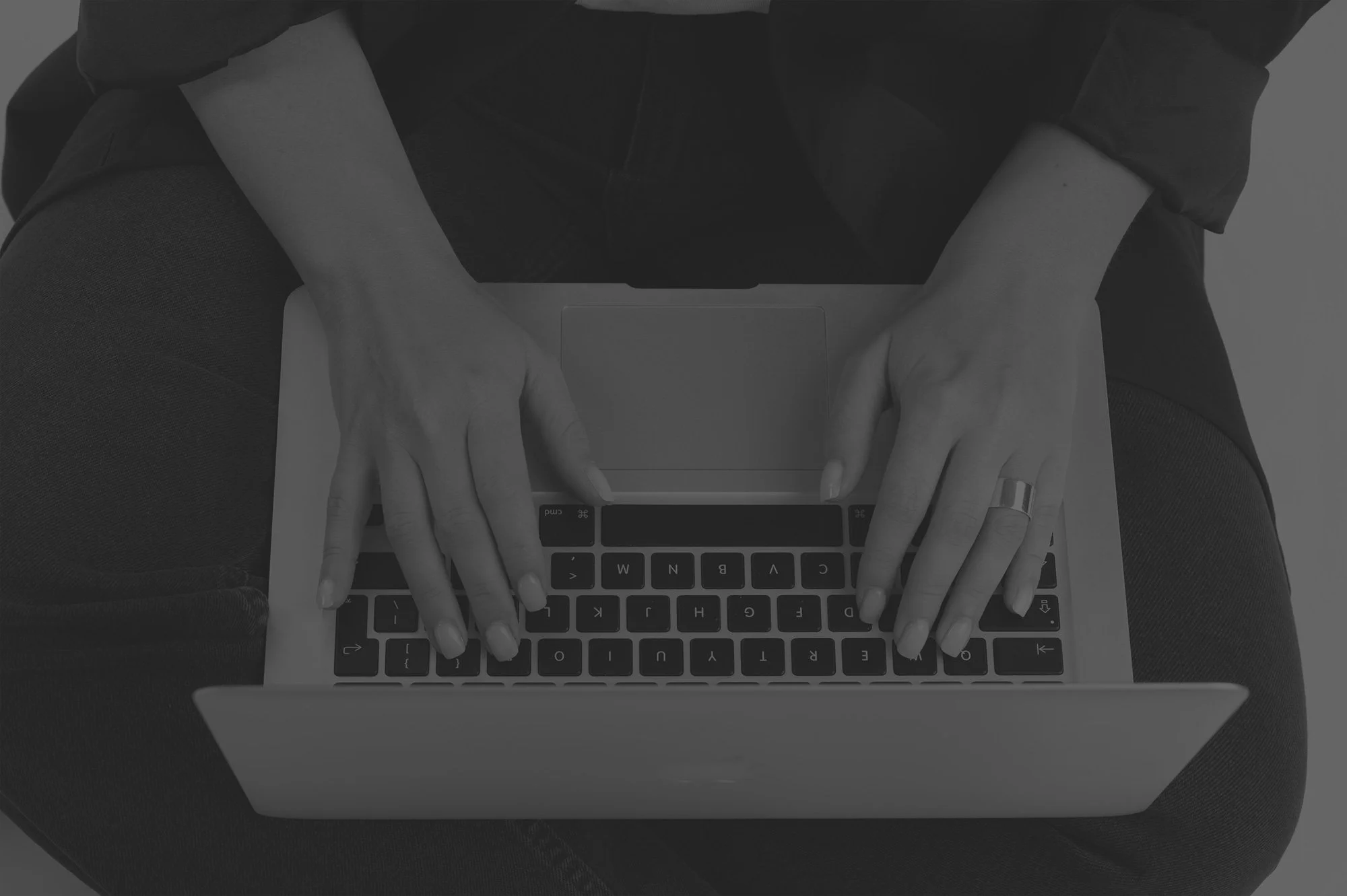Top-down view of a person wearing a dark long sleeve shirt and jeans, sitting cross-legged, typing on a MacBook laptop.