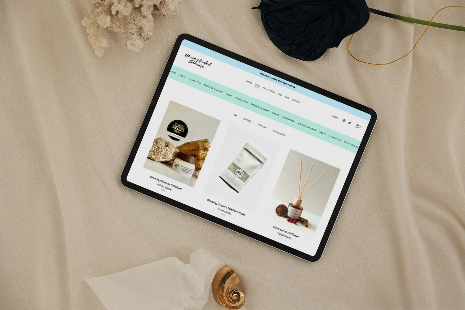 Tablet on beige fabric showing an online store with self-care and skincare products, with decorative items and a piece of crumpled tissue nearby.