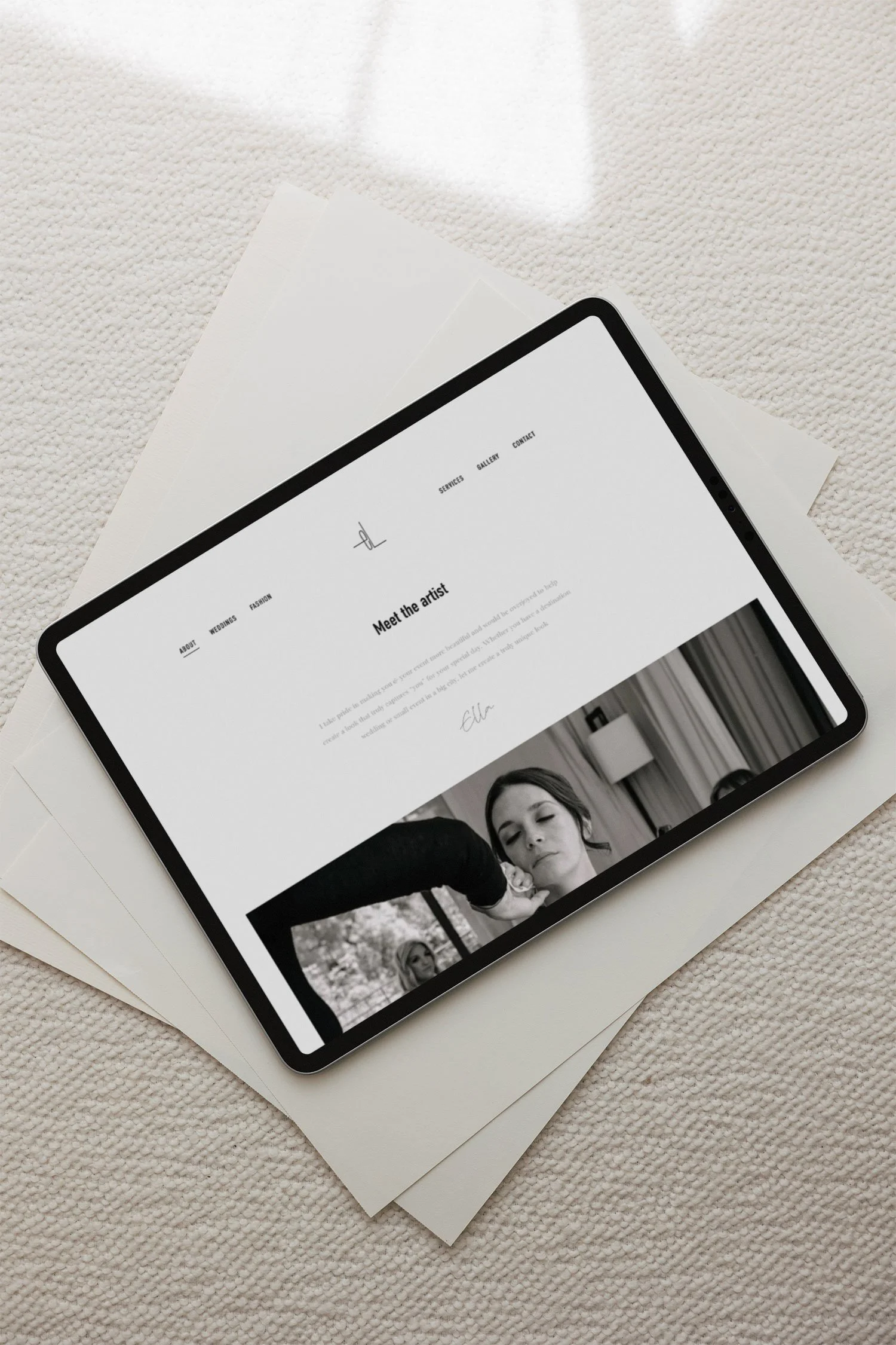 A tablet displaying a website titled 'Meet the artist' with a black and white photograph of a woman, resting on white sheets of paper on a textured surface.