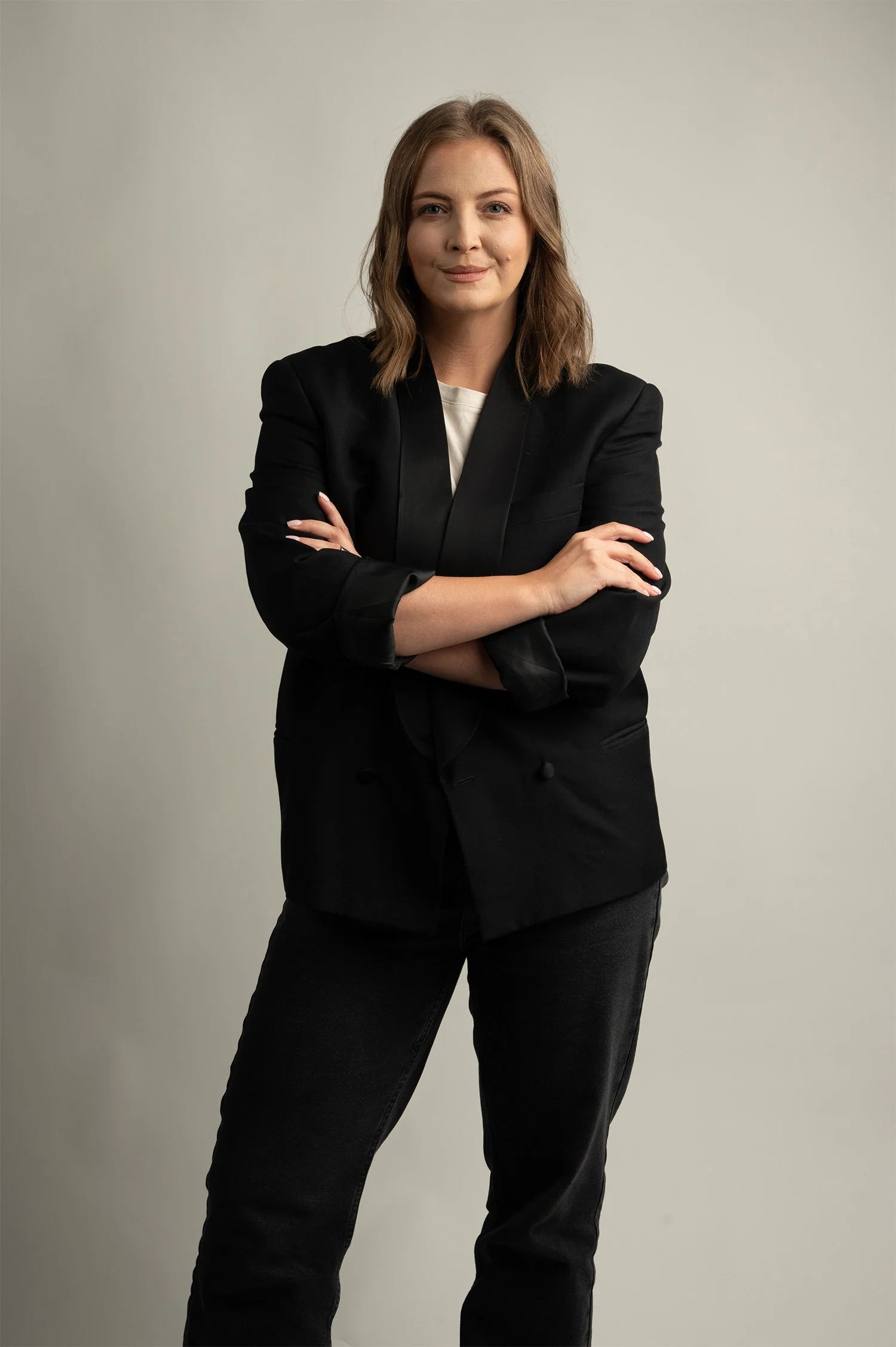 A woman with shoulder-length light brown hair, wearing a black blazer and black pants, standing against a plain light grey background with her arms crossed.