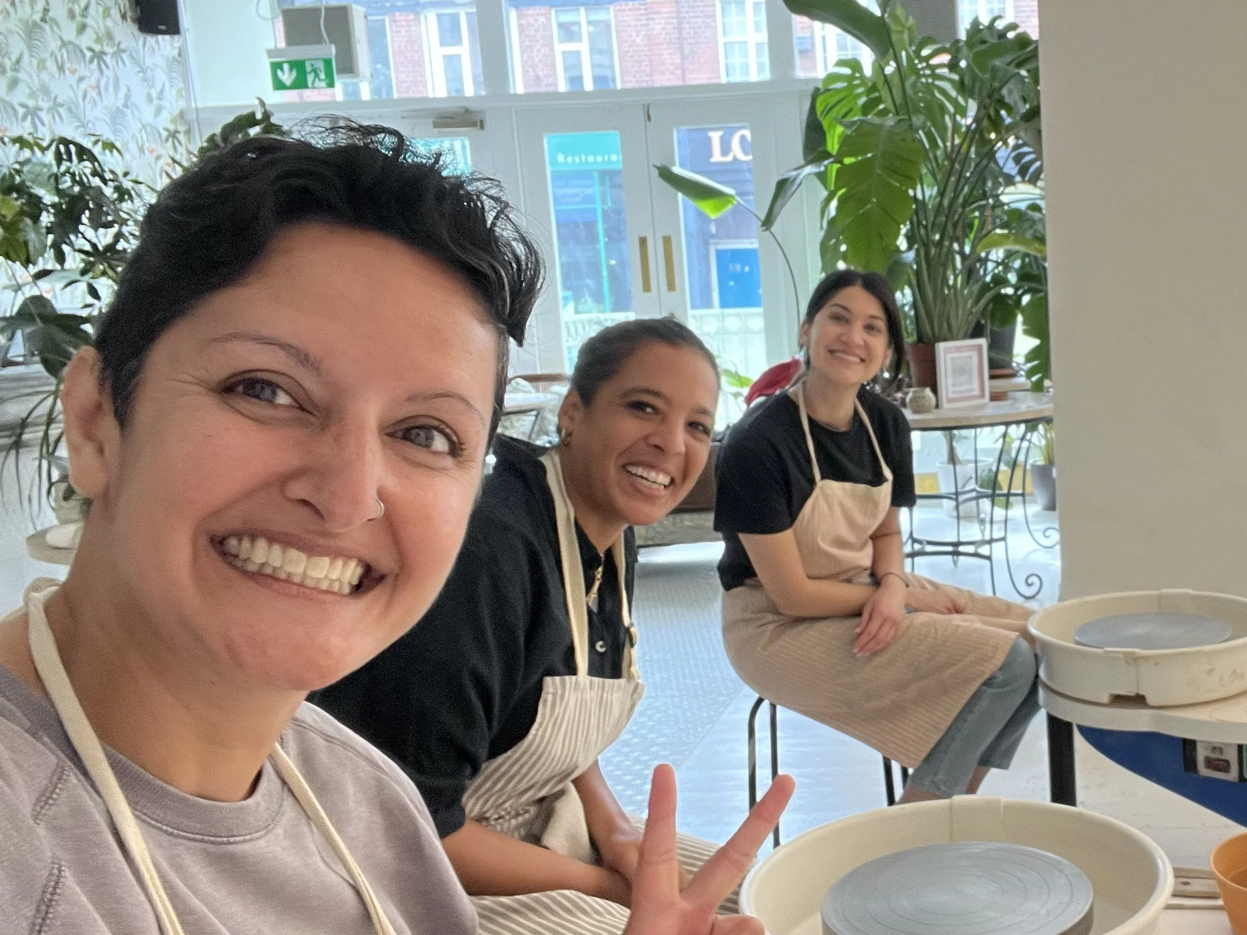 Four women sitting at a table with large bowls in front of them, smiling and making peace signs in a bright indoor space with green plants and large windows behind them.