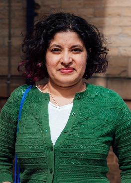 A woman with shoulder-length black curly hair with purple tips, wearing a green cardigan over a white shirt, standing outdoors in front of a wooden background.