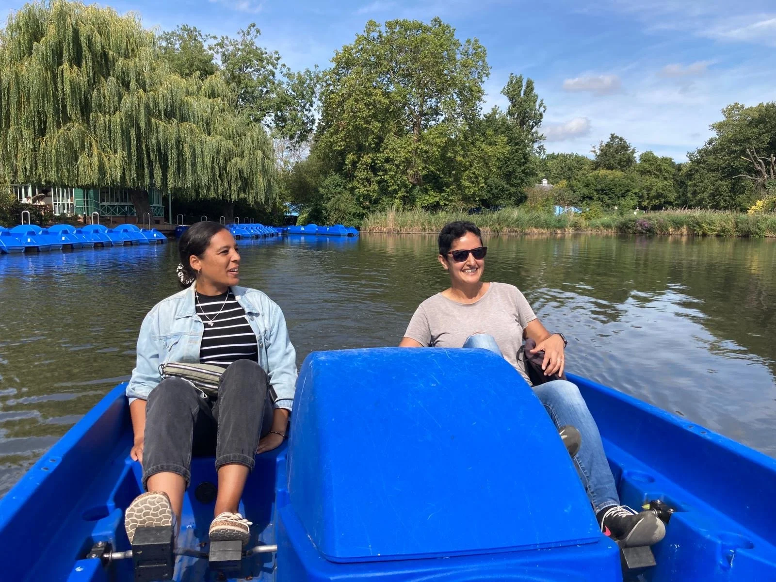 Two women are smiling and relaxing on a boat ride across a calm river with lush green trees in the background on a sunny day.