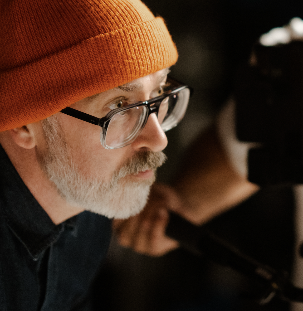 A middle-aged white man with an orange hat, gray beard, and glasses looking into a film camera. He wears a black shirt.