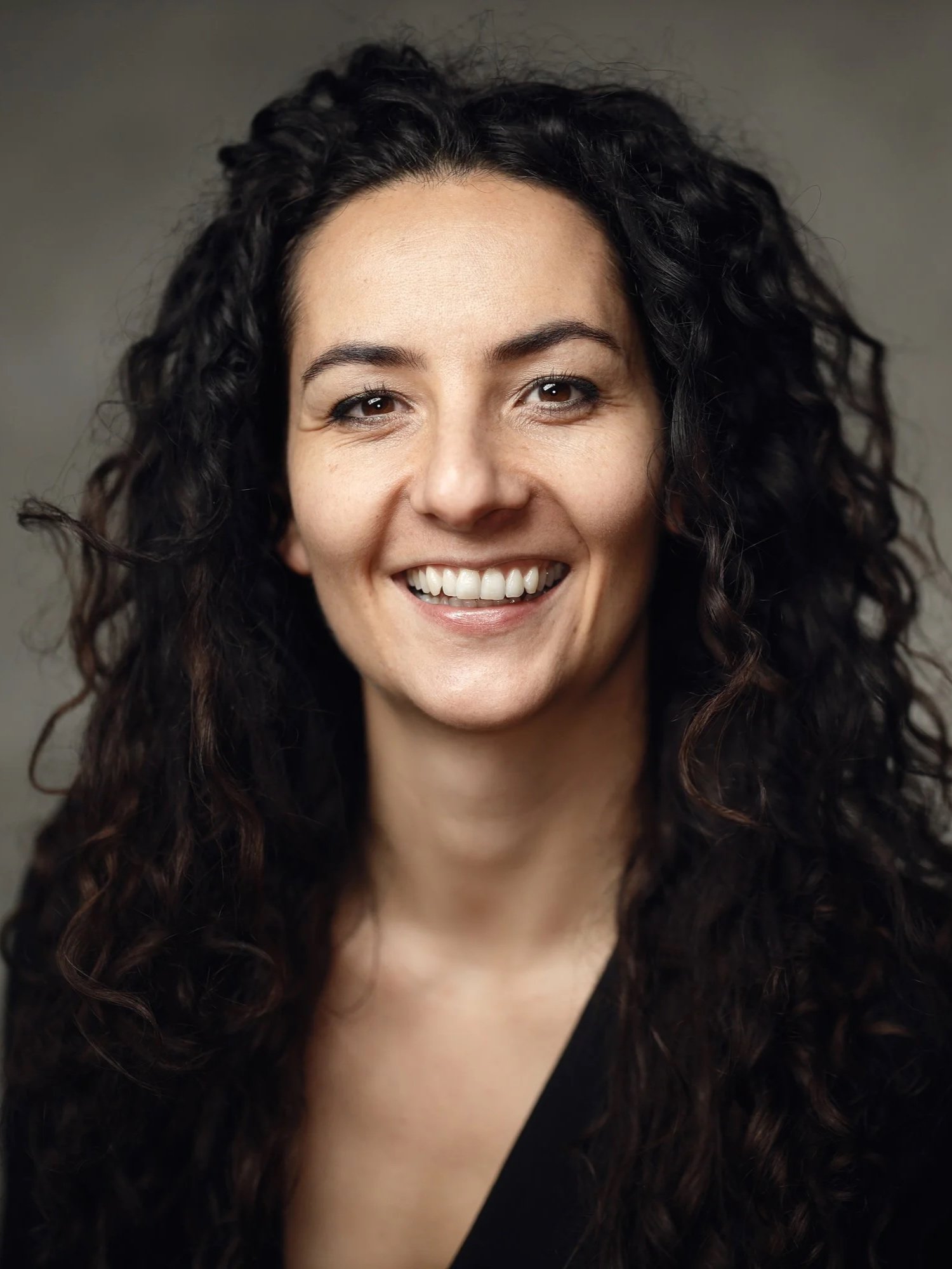A meditteraean white woman with long curly dark brown hair wearing a black top. She is smiling at the camera.