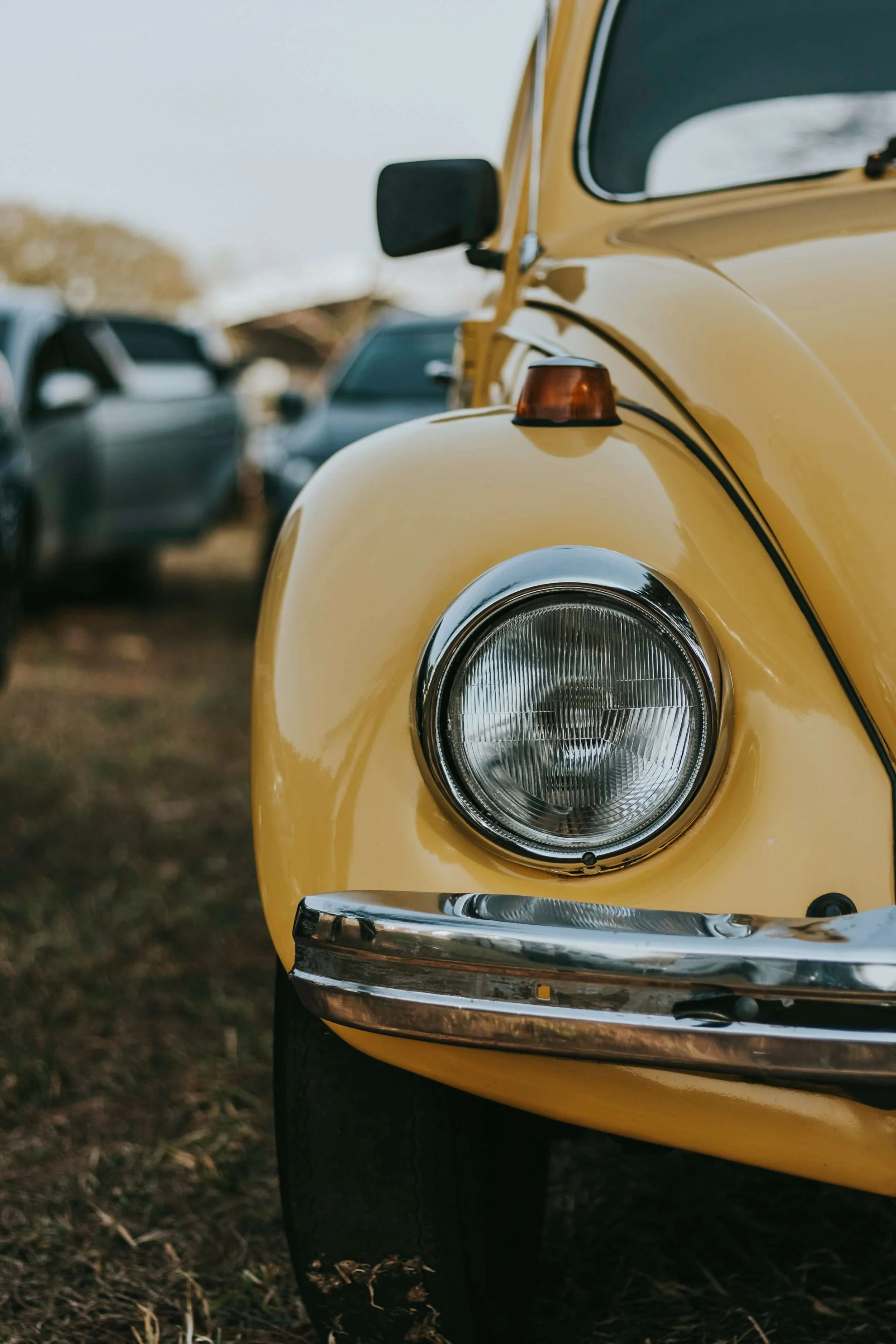 A close-up of the front of a yellow vintage car with a round headlight and a chrome bumper, parked outdoors on a dirt or grass surface with other cars in the background.