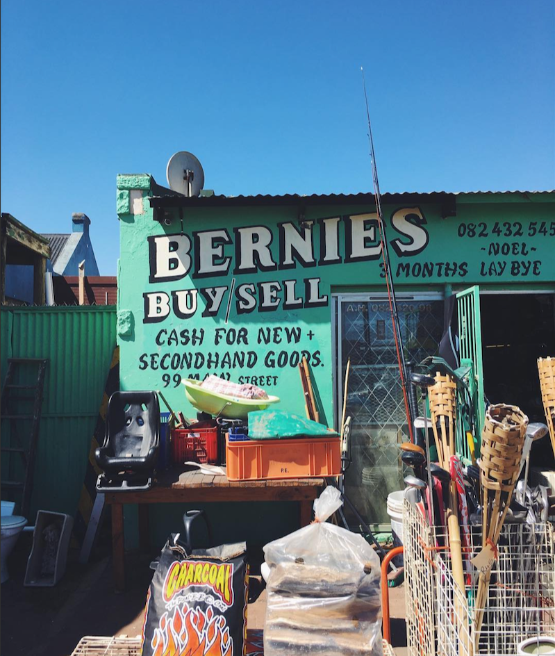 A photo of a small storefront with a green painted wall advertising Bernie's Buy and Sell, a thrift shop that buys and sells new and secondhand goods. The wall has a phone number and a sign that says '3 months laybye.' Outside, there are various items including a fishing rod, a plastic container with a boat-shaped object, wooden slats, and a bag of charcoal.