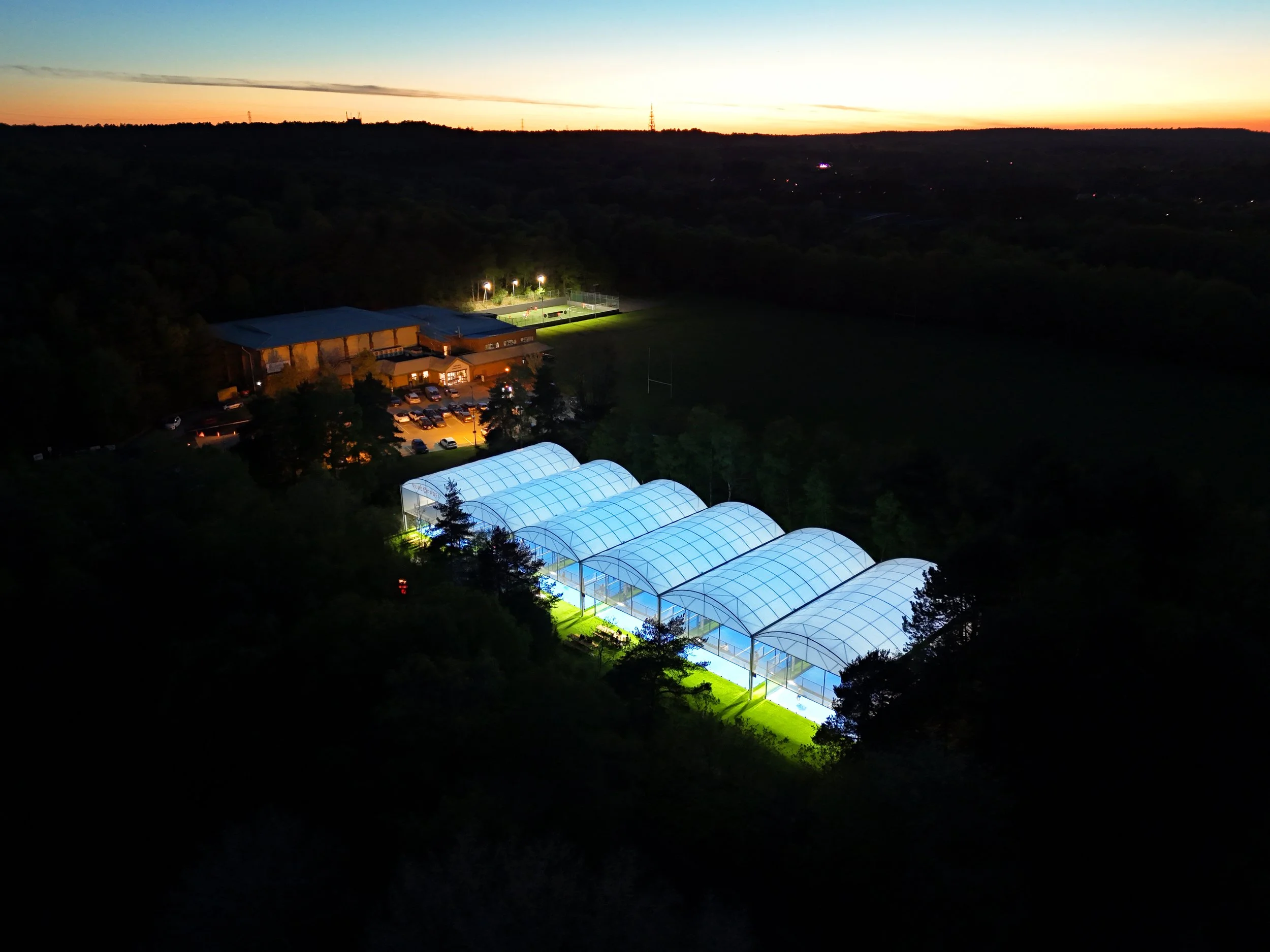 Aerial view of illuminated indoor padel courts at dusk surrounded by woodland and sports fields.