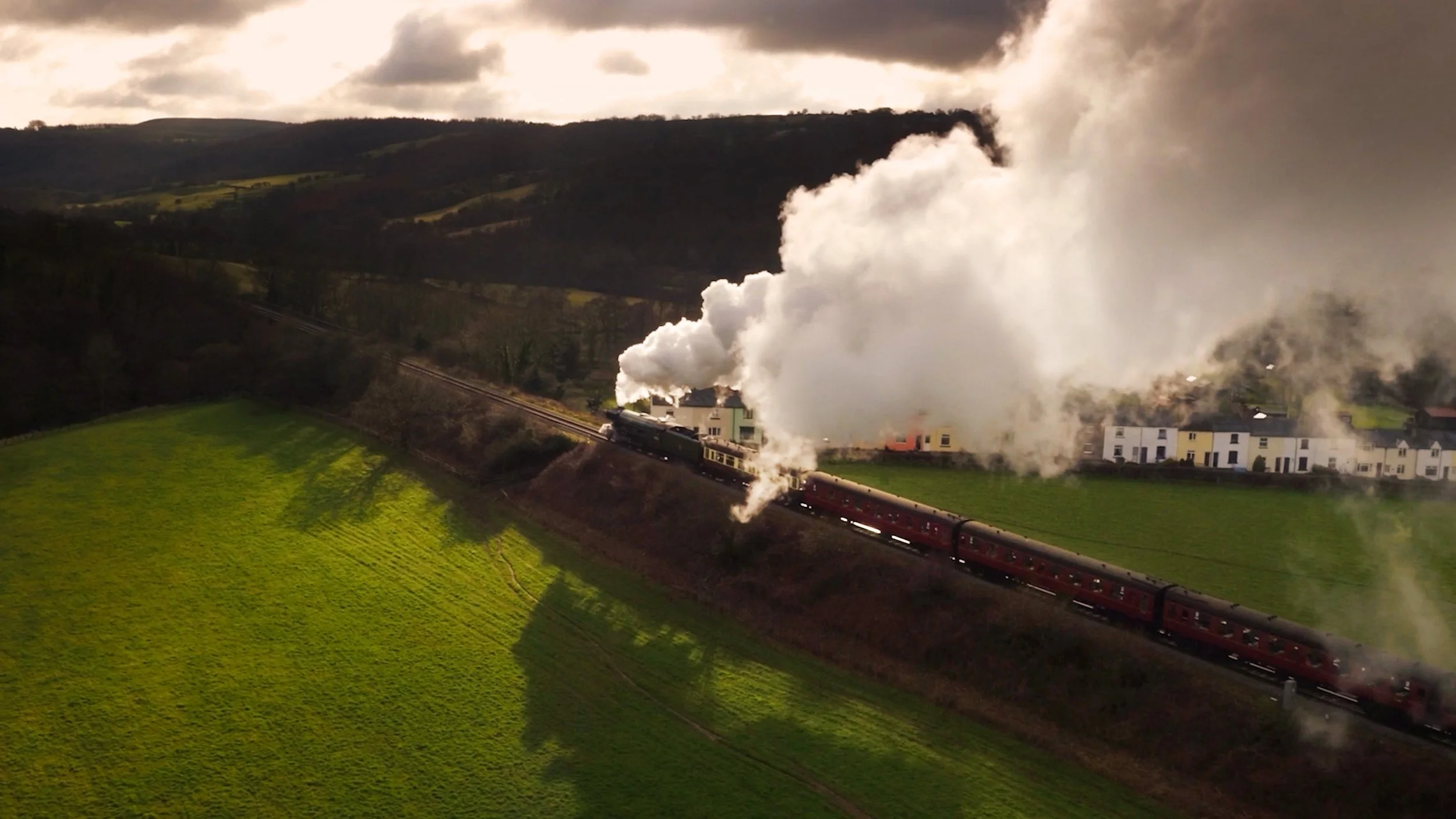 Aerial drone view of the Flying Scotsman steam locomotive travelling through the British countryside, with billowing steam and the historic train cutting through the landscape.