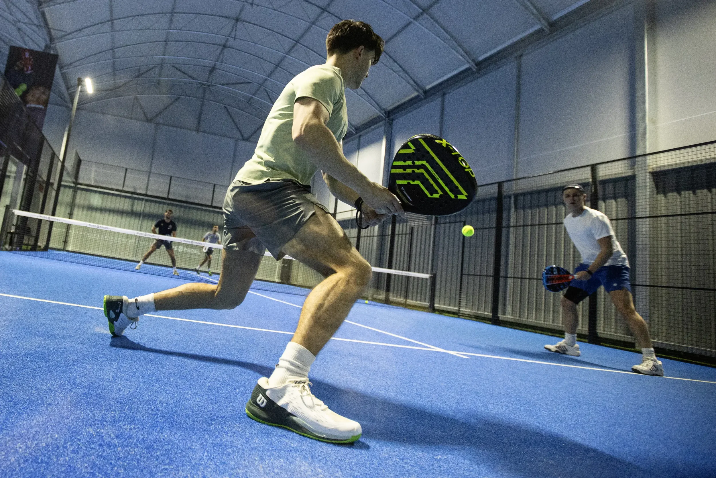 Photograph by Lewis Whyld of a Padel player lunging forward to return a low ball during a doubles rally on an indoor blue court.