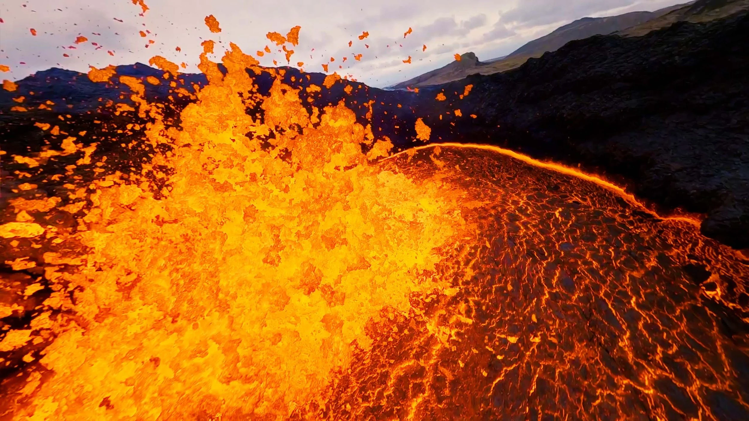 Aerial drone view of an active volcano in Iceland at sunset, with glowing lava, volcanic terrain and dramatic smoke rising across the rugged landscape.