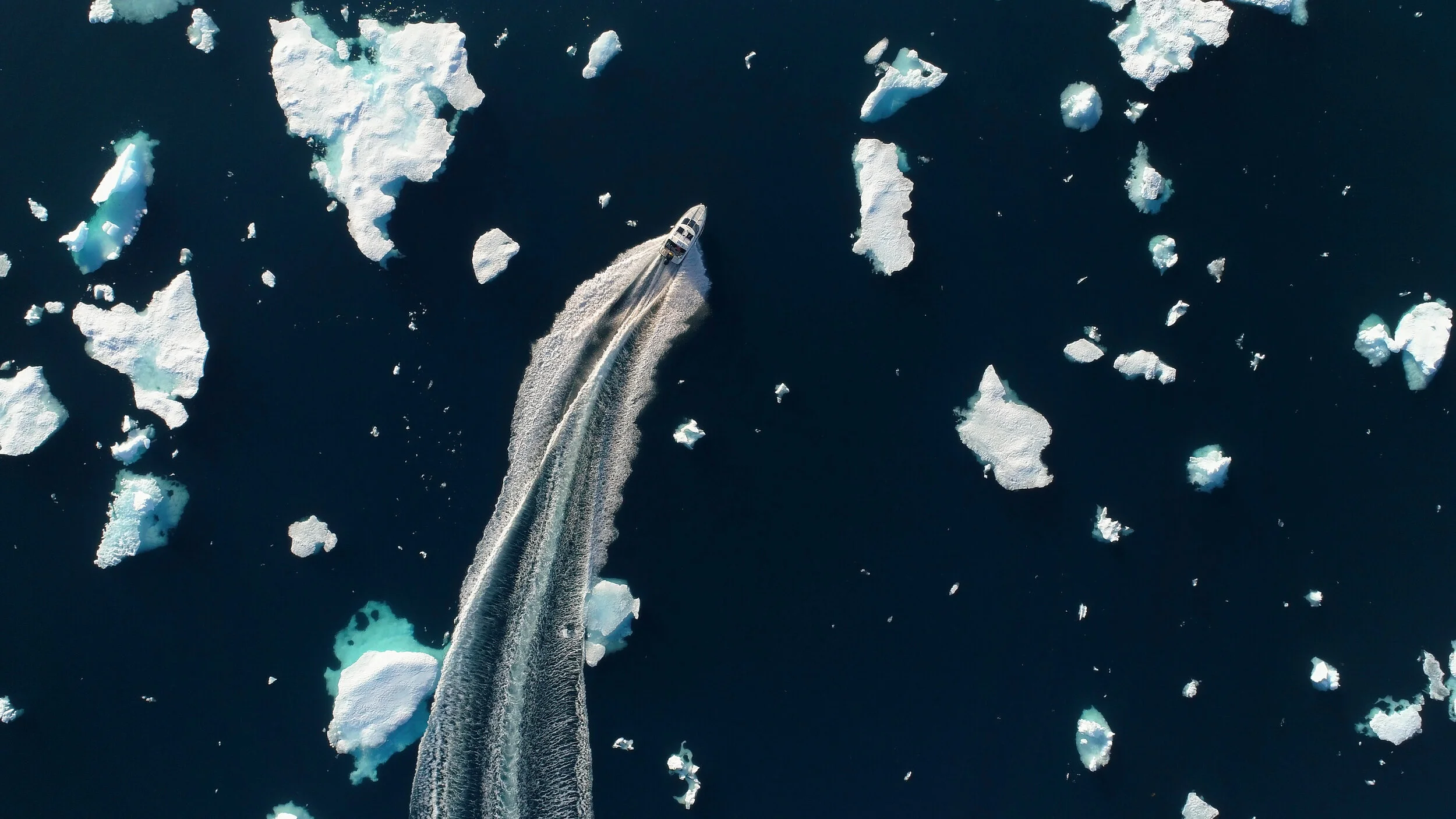 Aerial view of a boat travelling through sea ice on the journey to Summit Station, surrounded by broken Arctic ice and the vast frozen seascape of Greenland.