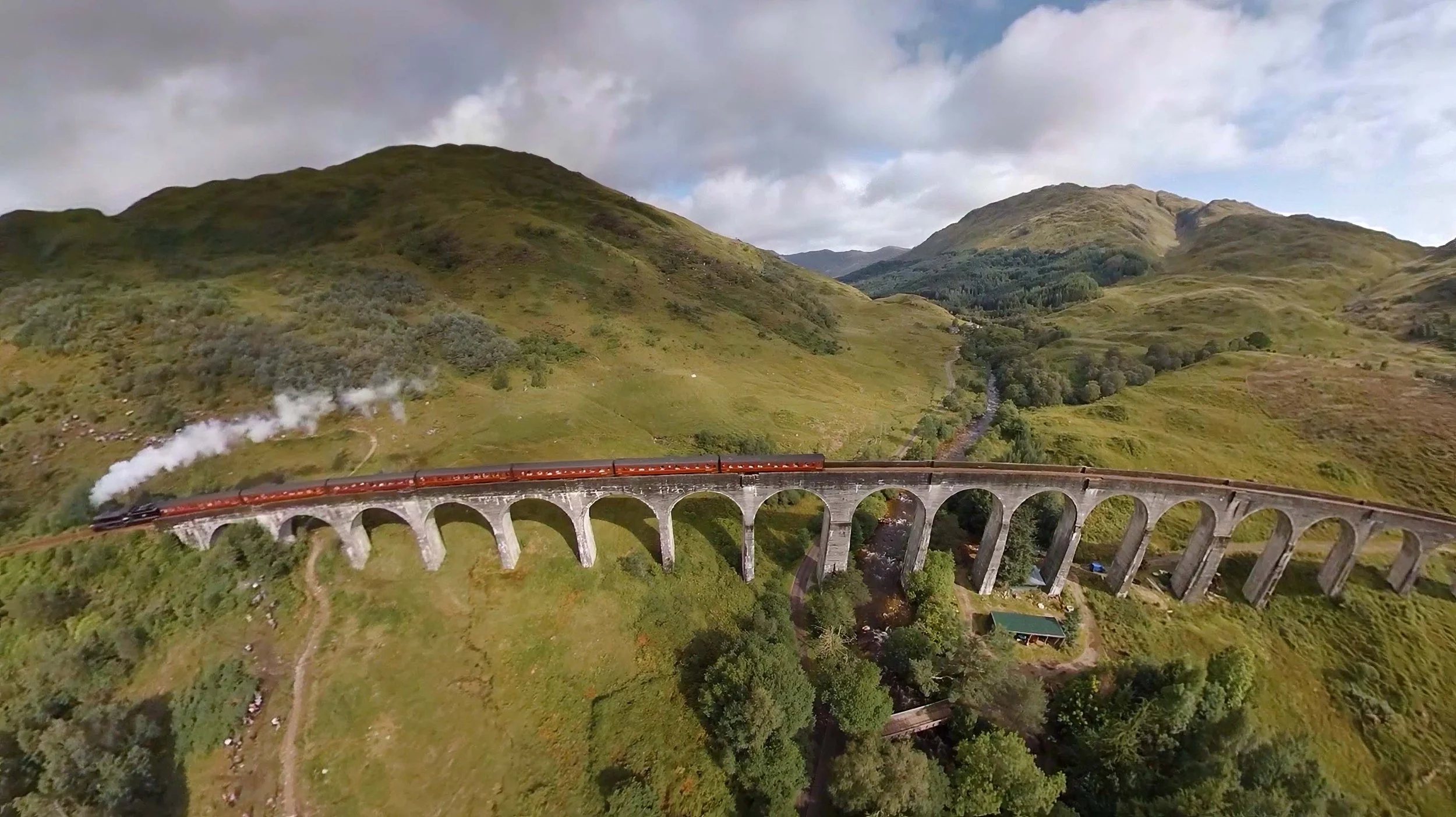 Aerial drone view of the Jacobite steam train, known as the “Hogwarts Express,” travelling through the Scottish Highlands with dramatic mountain scenery and historic railway landscape.