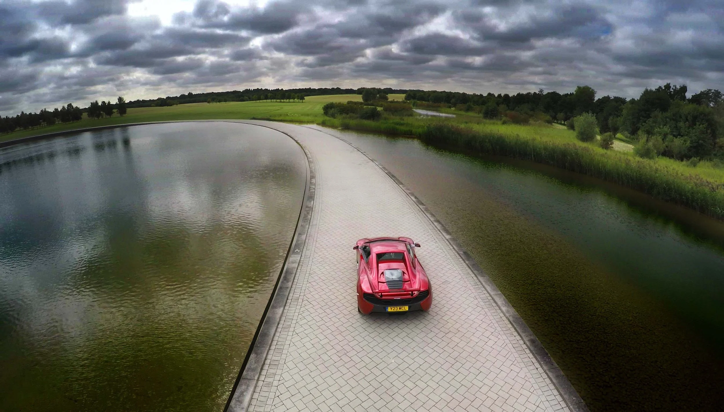 FPV drone view chasing a McLaren around the reflective water feature outside the McLaren Technology Centre, capturing the car in motion against the iconic curved glass headquarters.