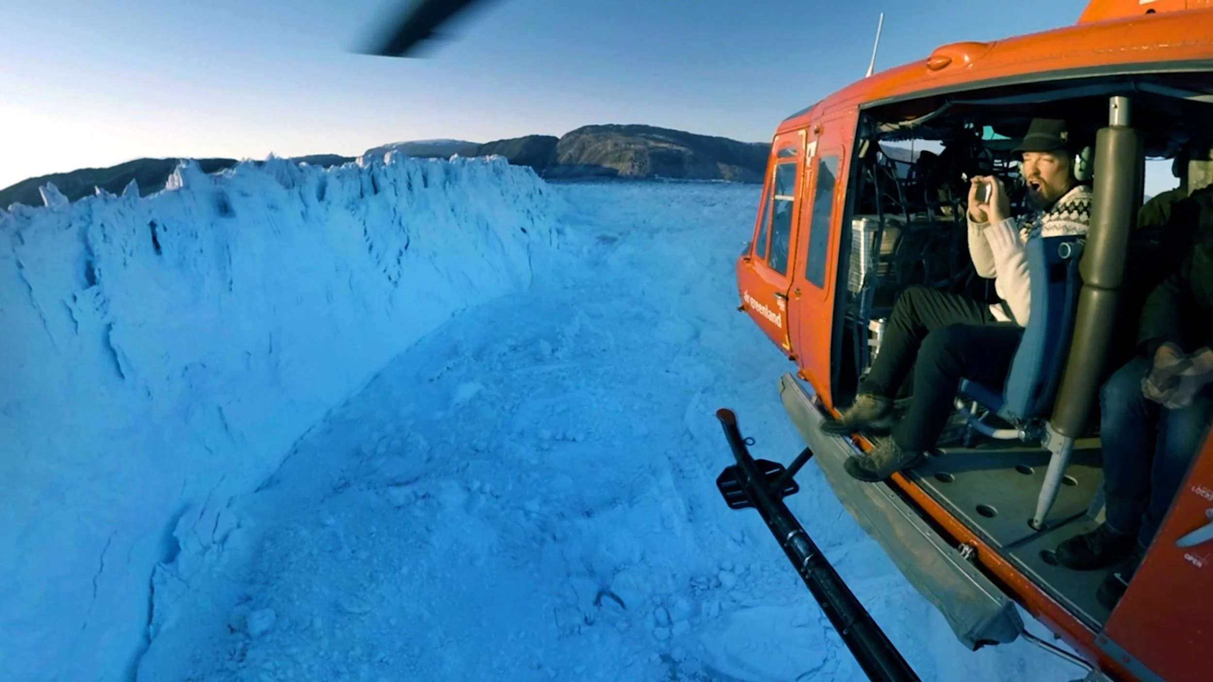 Dramatic aerial helicopter view over Helheim Glacier in Greenland at sunset, with golden light illuminating the vast ice sheet, crevasses and Arctic landscape.