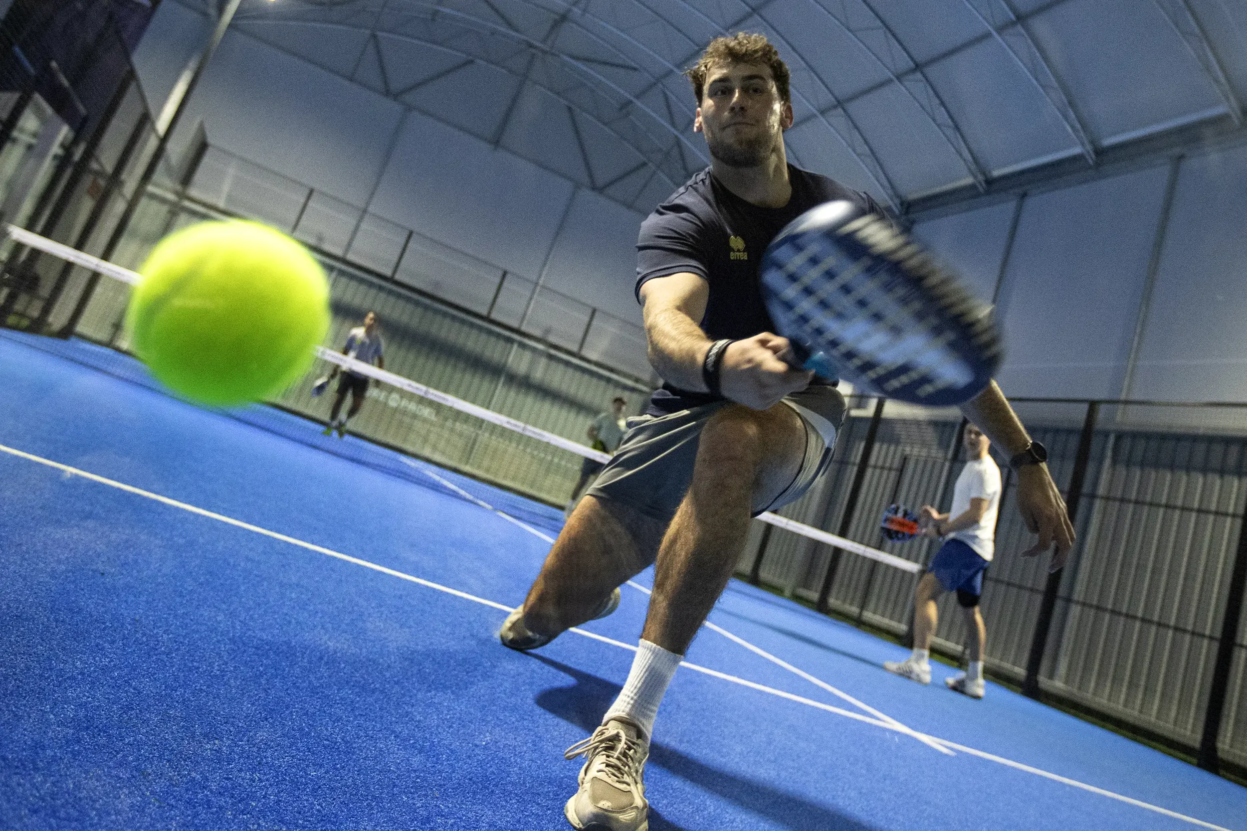 Padel player reaching for a fast ball close to the camera during an intense indoor doubles match. Photograph by Lewis Whyld