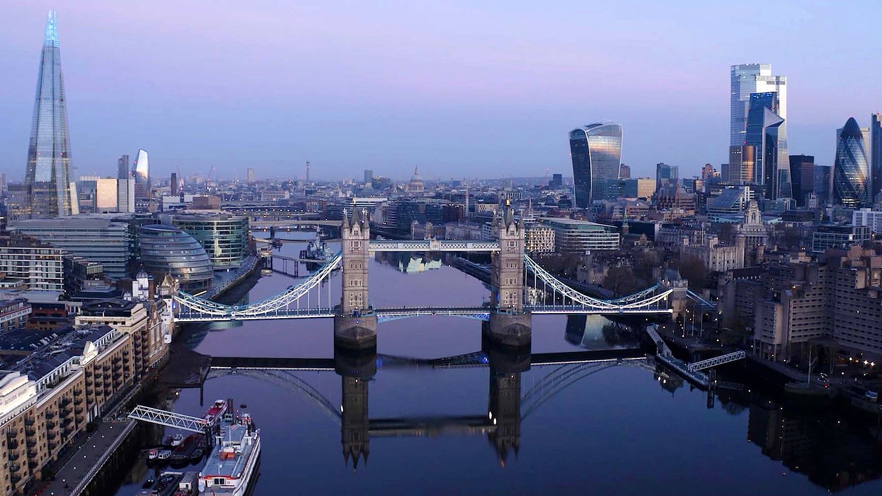 Aerial drone view of Tower Bridge at sunset, with warm golden light over the River Thames, the historic bascules and towers, and the London skyline beyond.