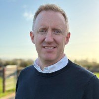 Portrait of a smiling man with short red hair and a beard, wearing a checked shirt and a navy vest, outdoors with a blurred green background.