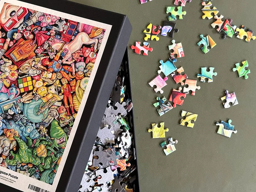 Colorful jigsaw puzzle about a cartoon scene with various characters and objects, partially assembled in a black box, with loose puzzle pieces scattered on a green surface.
