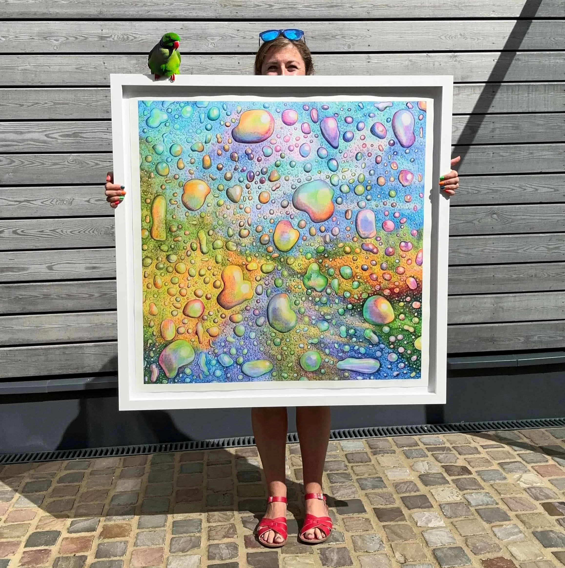 Gemma doodle is holding a large colorful abstract pencil drawing with water droplet patterns, standing outside on a cobblestone surface, with a wooden wall in the background.