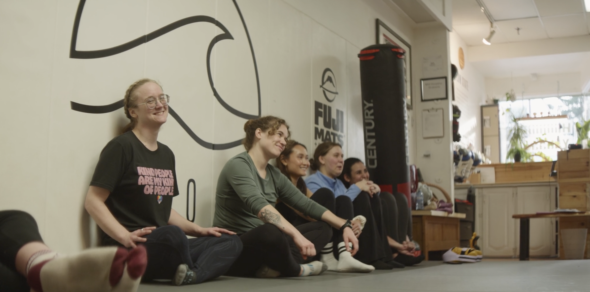 Group of five people sitting against a wall in a room, smiling and listening. The room has a large wave logo on the wall and shelves with decorations, plants, and shoes.