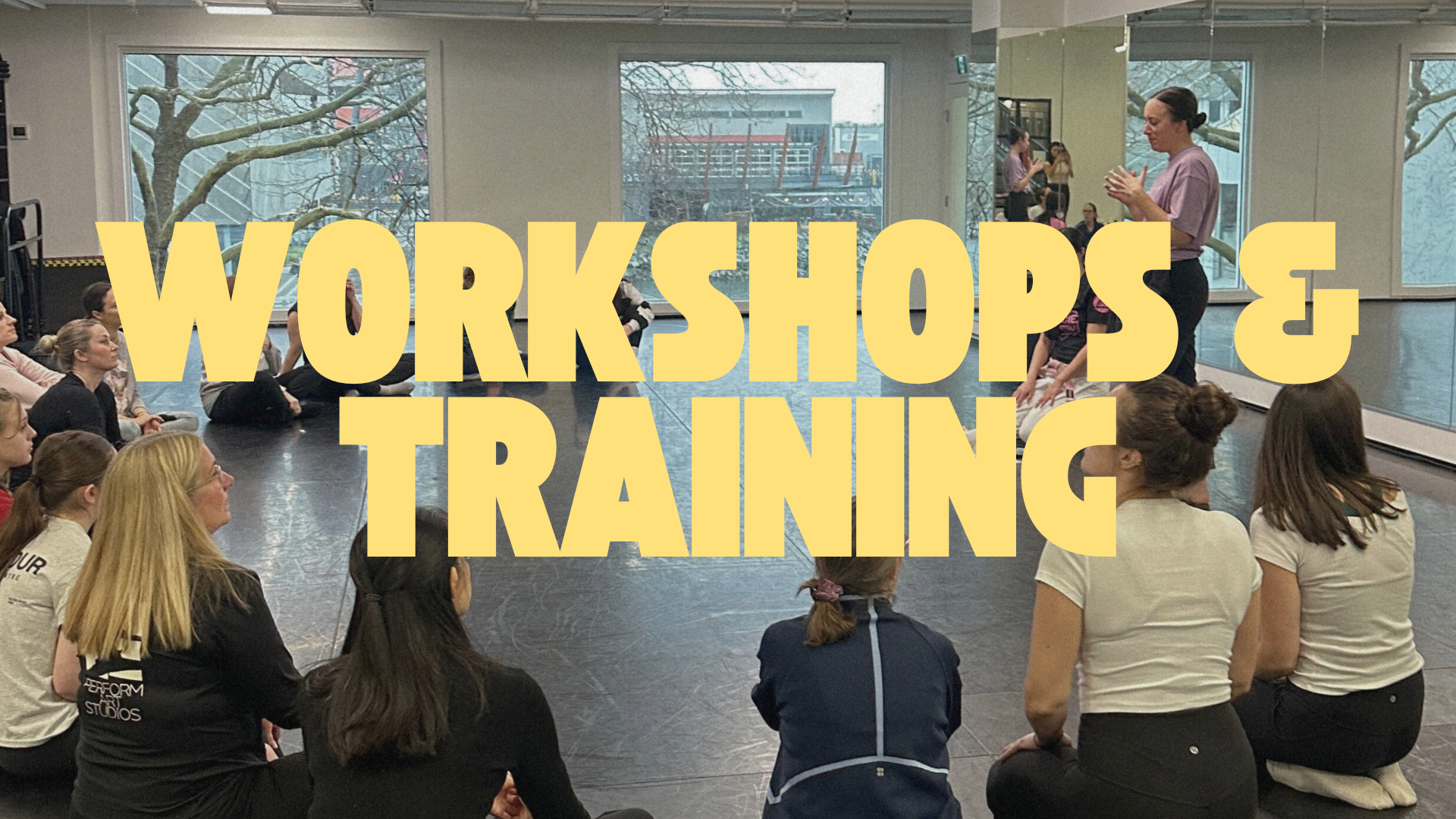 A group of people sitting on the floor of a studio, listening to a woman standing and speaking at the front. The scene appears to be a workshop or training session, with the words 'Workshops and Training' in large yellow text overlaying the image.