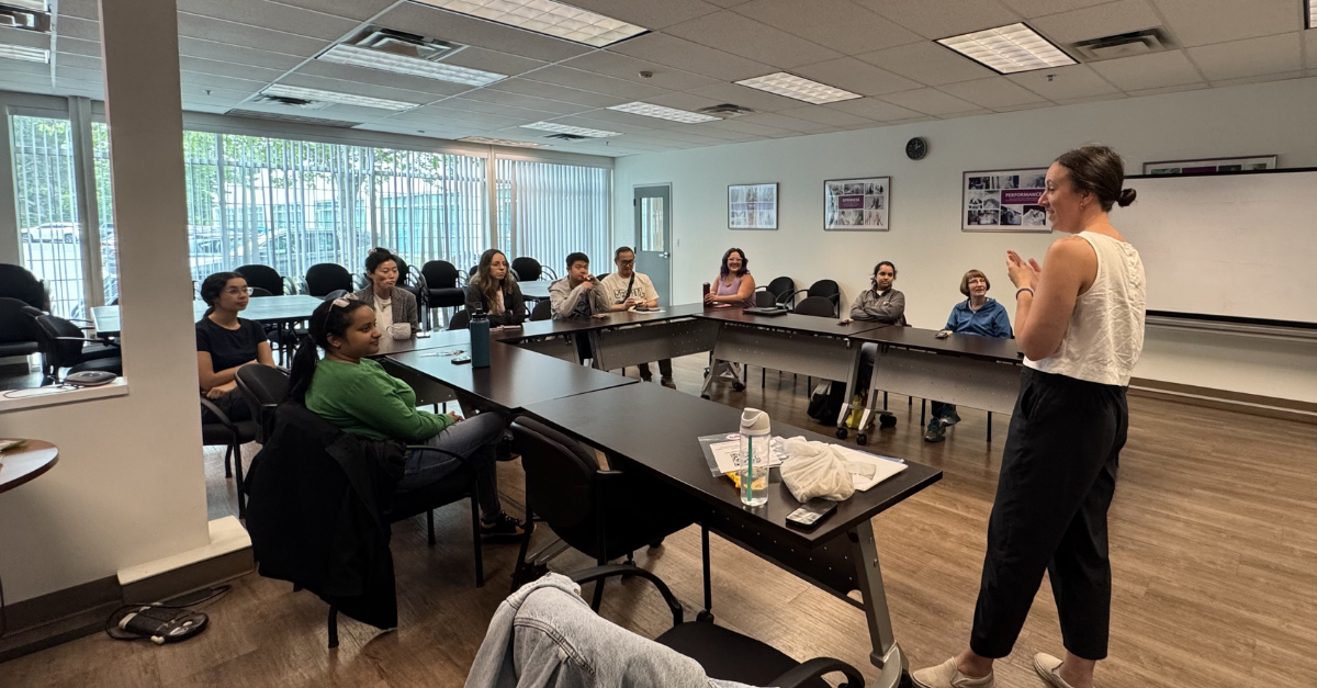 A woman giving a presentation to a group of people seated around a U-shaped conference table in a room with large windows and framed posters on the wall.