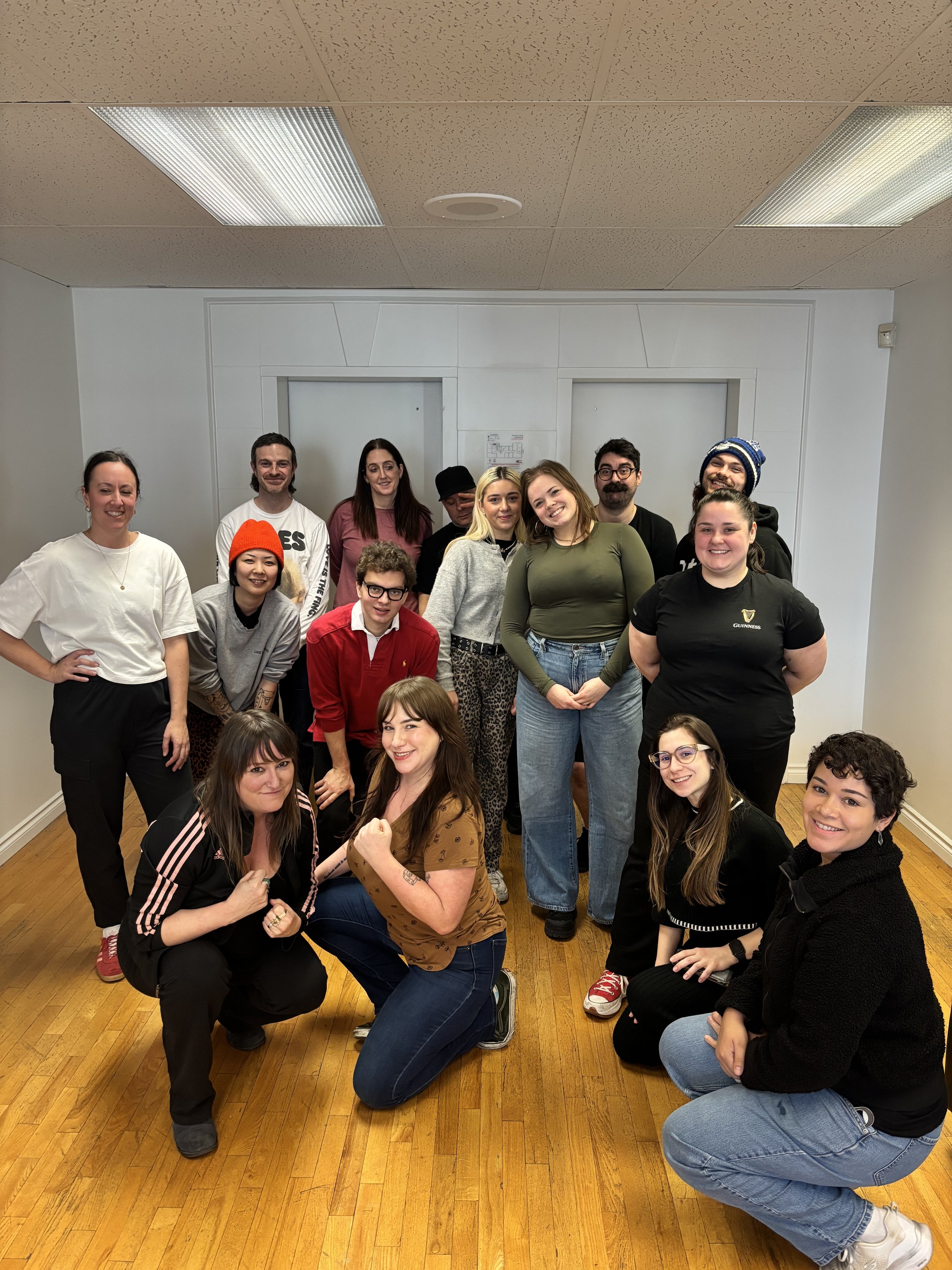 A group of diverse young adults gathered in a room with wooden floors, posing for a photo. Some are standing, some are kneeling or squatting, smiling and showing various expressions of camaraderie.