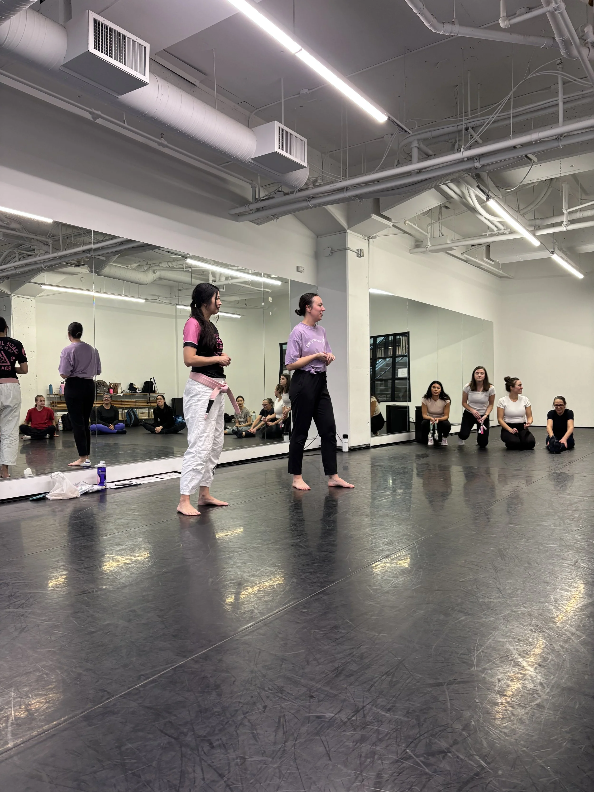 Group of women standing and sitting in a dance studio with a large mirror, black floor, and white walls. Two women are standing in the center, one with a pink shirt and one with a lavender shirt, barefoot. Several women are sitting against the wall,