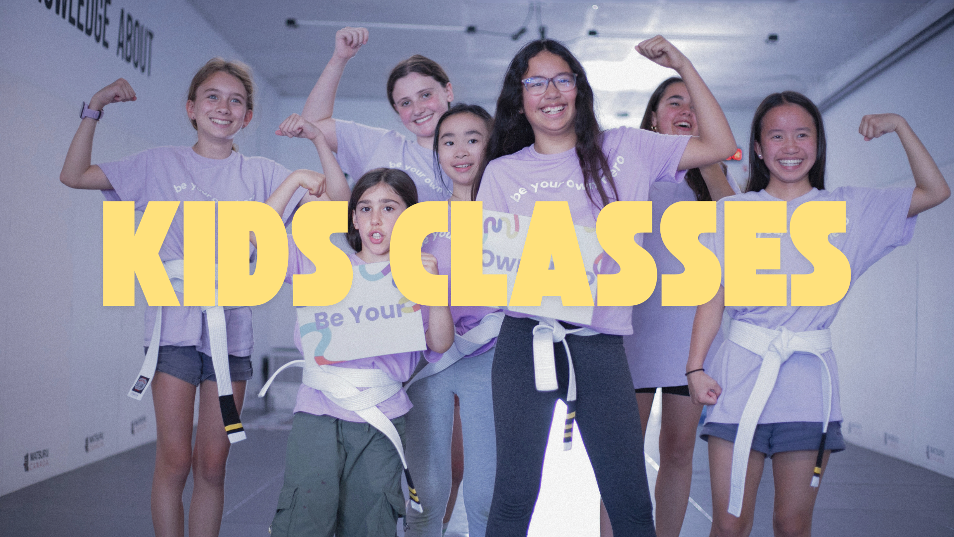 A group of seven young girls wearing purple t-shirts and white belts, striking a triumphant pose in a studio, with the words 'Kids Classes' in large yellow text overlaying the image.