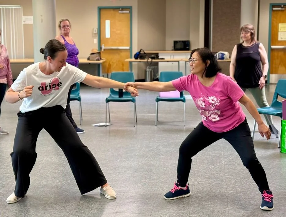 Two women engaged in a playful stretching or dance activity in a community room, with others watching in the background.